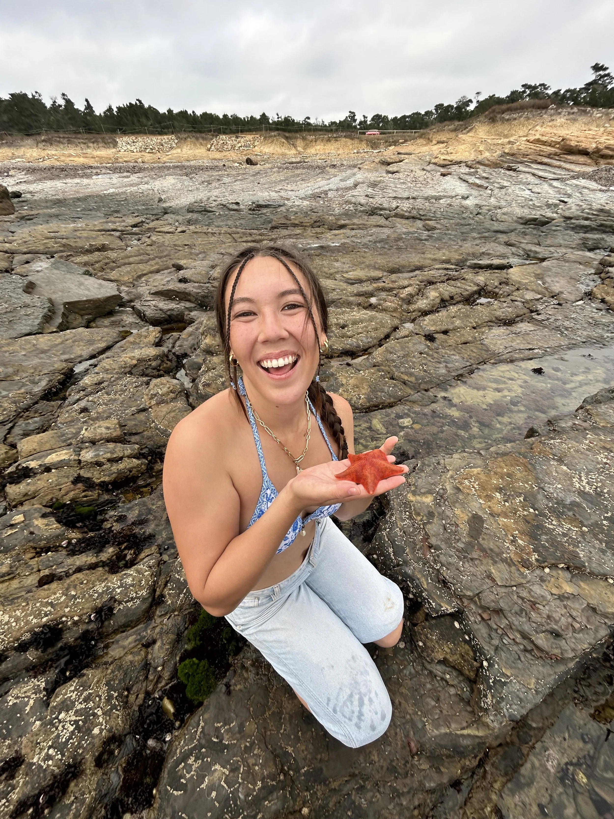 A young woman with braided hair, smiling and holding a red starfish while kneeling on rocks near the water at the beach.