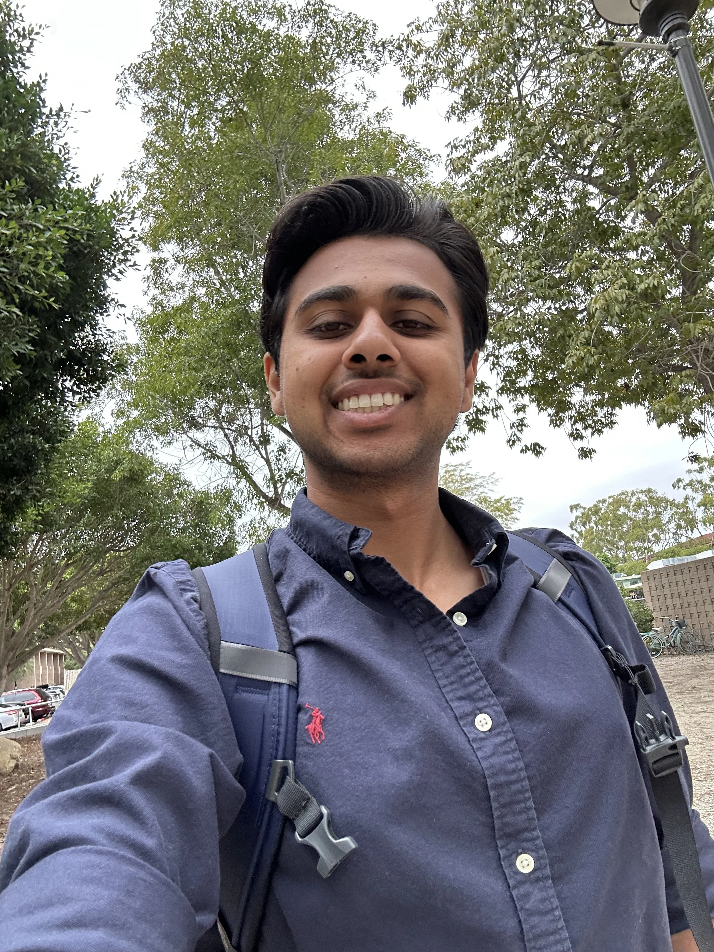 A young man taking a selfie outdoors, smiling, wearing a dark blue button-up shirt and carrying a backpack, with trees and cars in the background.