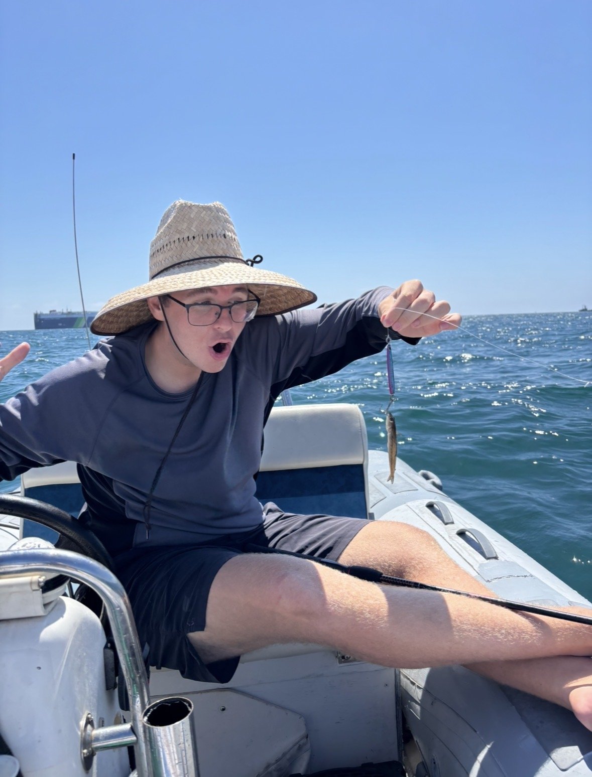 Young man on a boat, wearing a wide-brimmed hat and glasses, holding a small fish on a fishing line with a surprised expression.