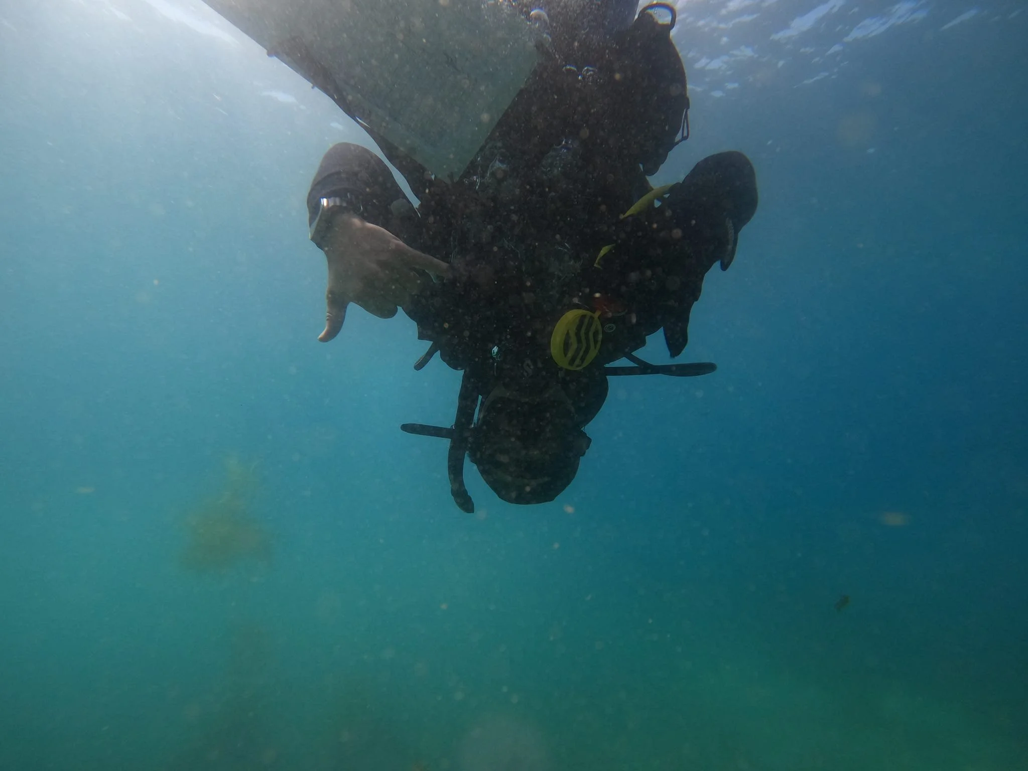Scuba diver underwater making an 'OK' hand signal, with sediment and water particles surrounding them and a faint blurry object in the distance.