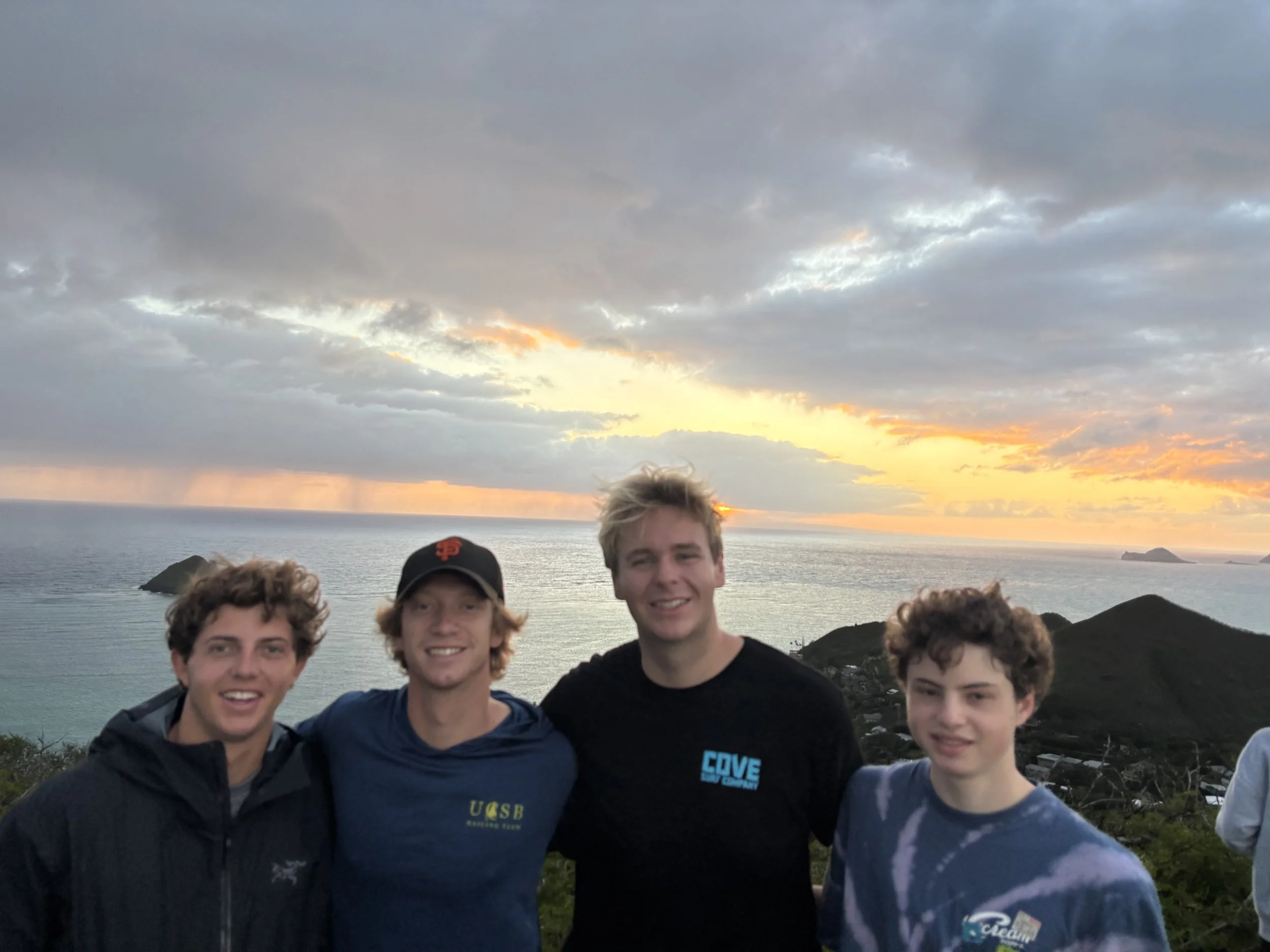 Four young men smiling at sunset on a hill overlooking the ocean with small islands in the background.