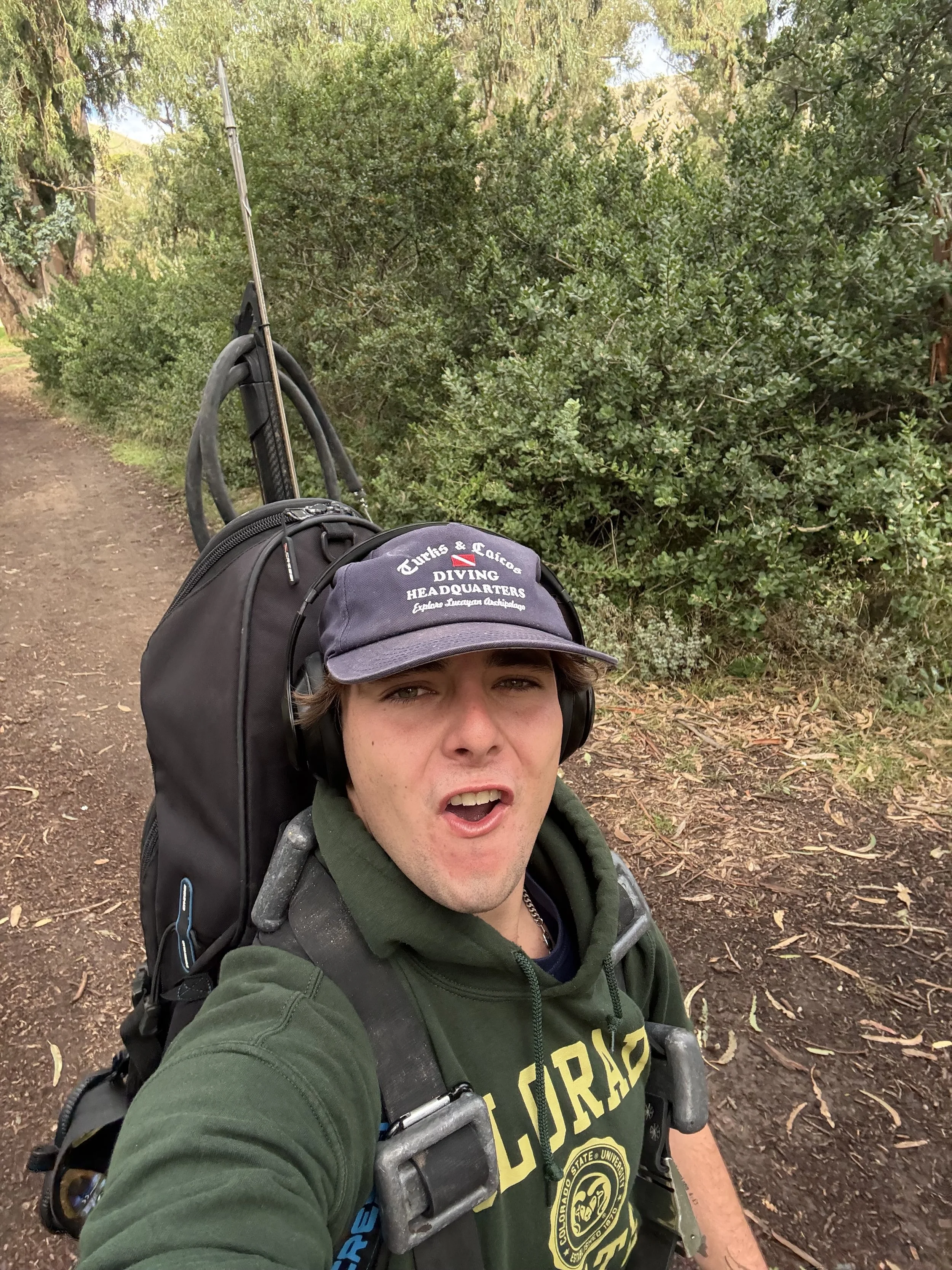 A young man taking a selfie outdoors on a trail, wearing a hat, headphones, a green hoodie, and carrying a backpack with diving gear straps, surrounded by trees and bushes.