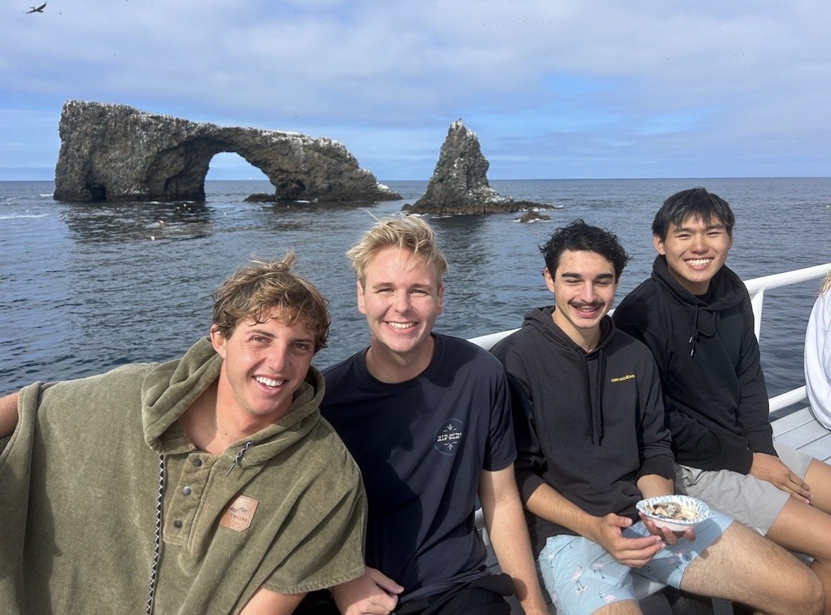 Four young men smiling and sitting on a boat with ocean and rock formations in the background.