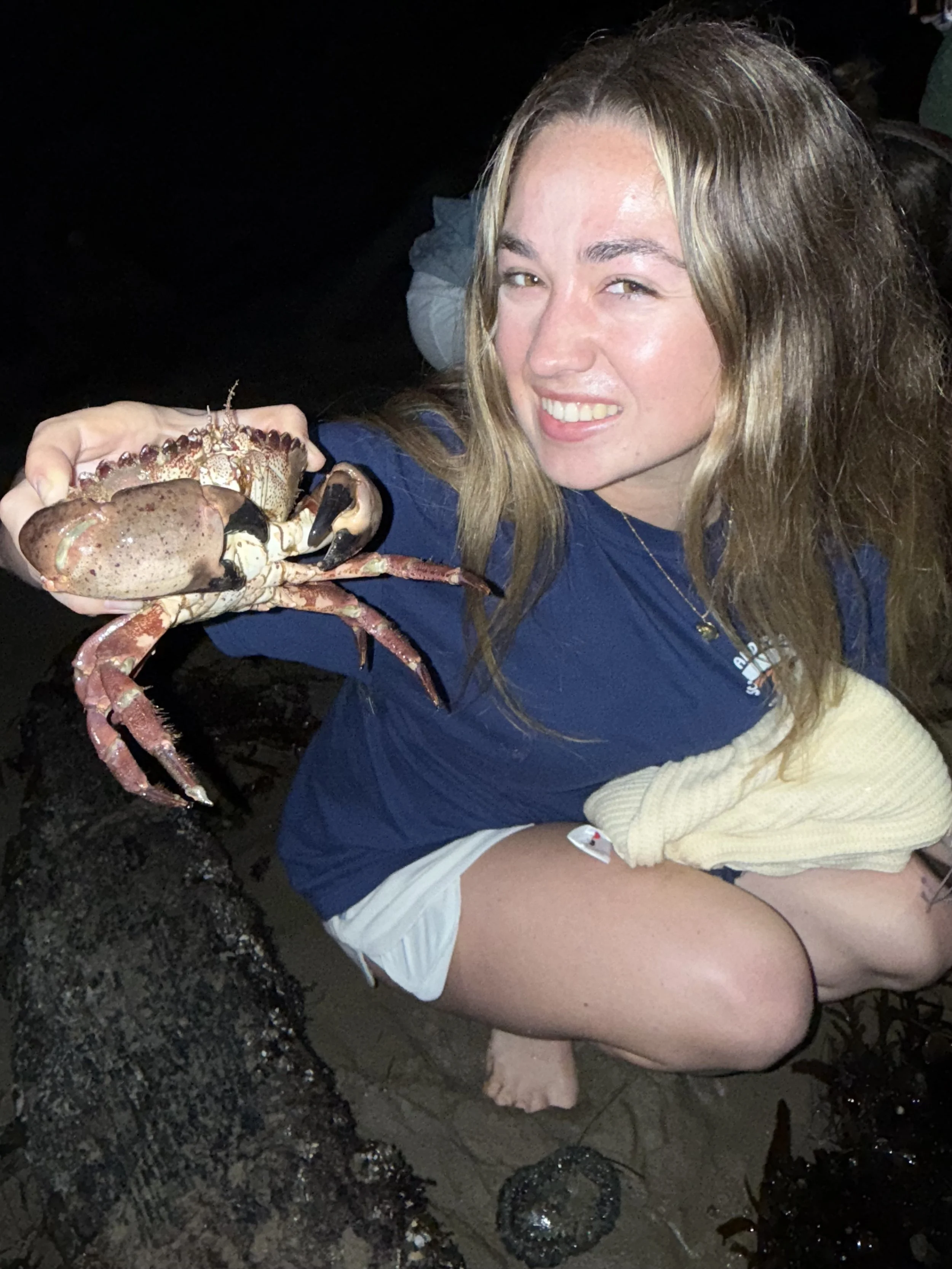 A woman smiling and crouching on the sand at night, holding a large crab in her hand.