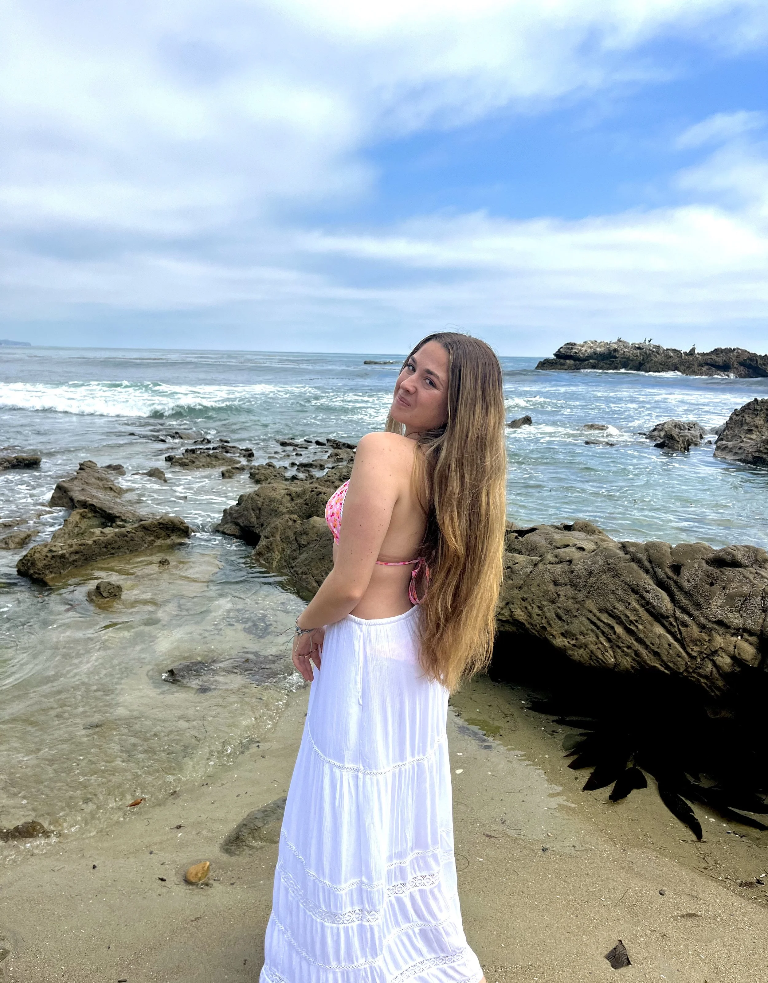 Young woman standing on a sandy beach with rocks, ocean waves, and a partly cloudy sky in the background.