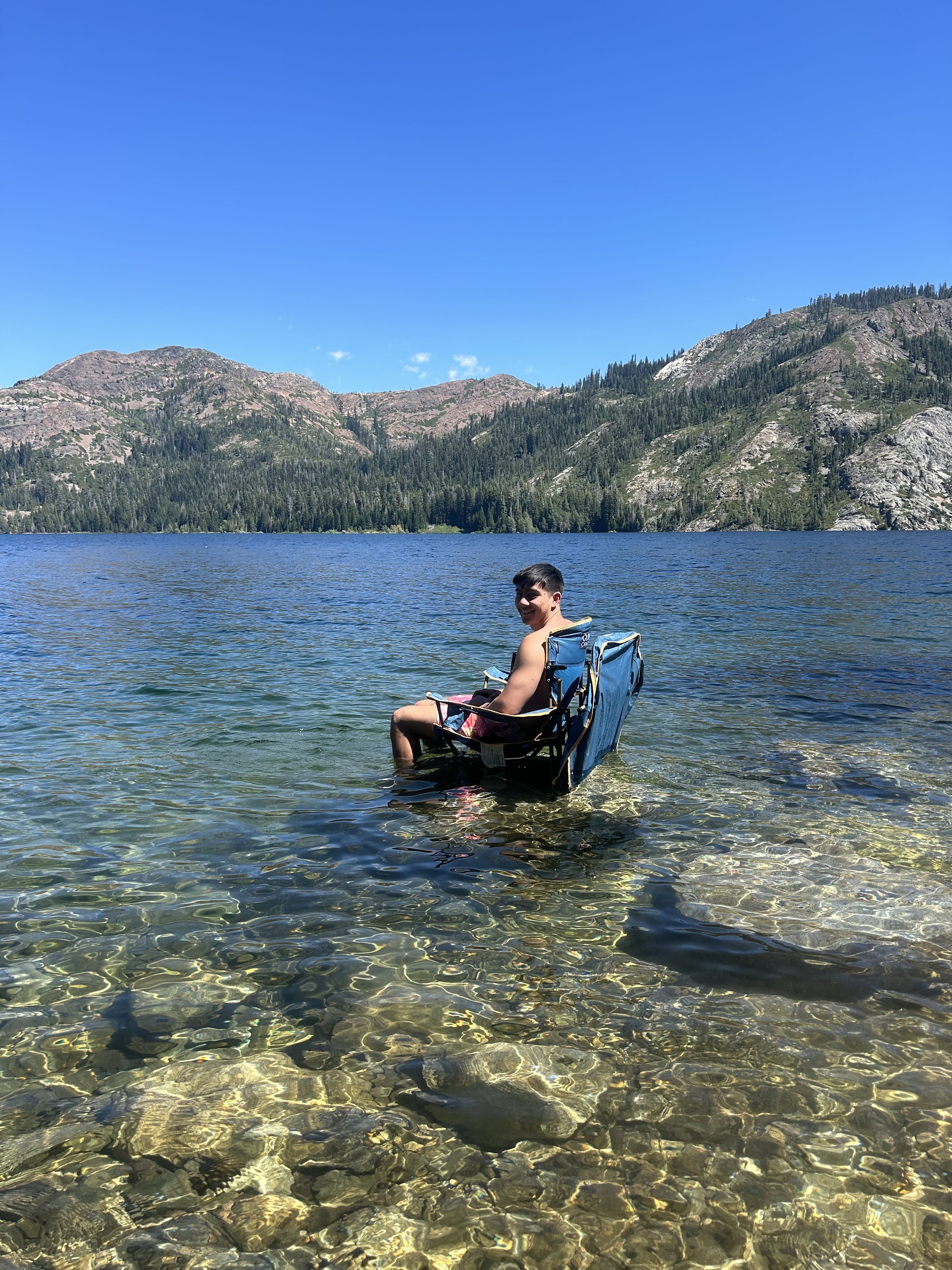 A person sitting in a blue camping chair in clear water at a lake with mountainous terrain and pine trees in the background under a blue sky.