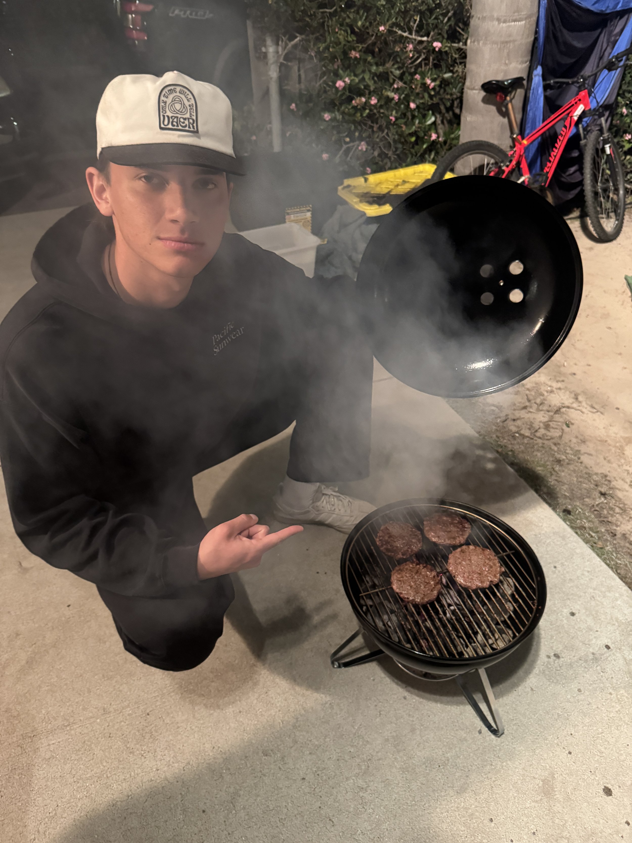 A young man kneeling outside at night, holding a black lid over a small charcoal grill with four patties cooking on it. The man is wearing a white and black cap, a black hoodie, and white sneakers. In the background, there is a bicycle, a blue blanket, and some plants with pink flowers.