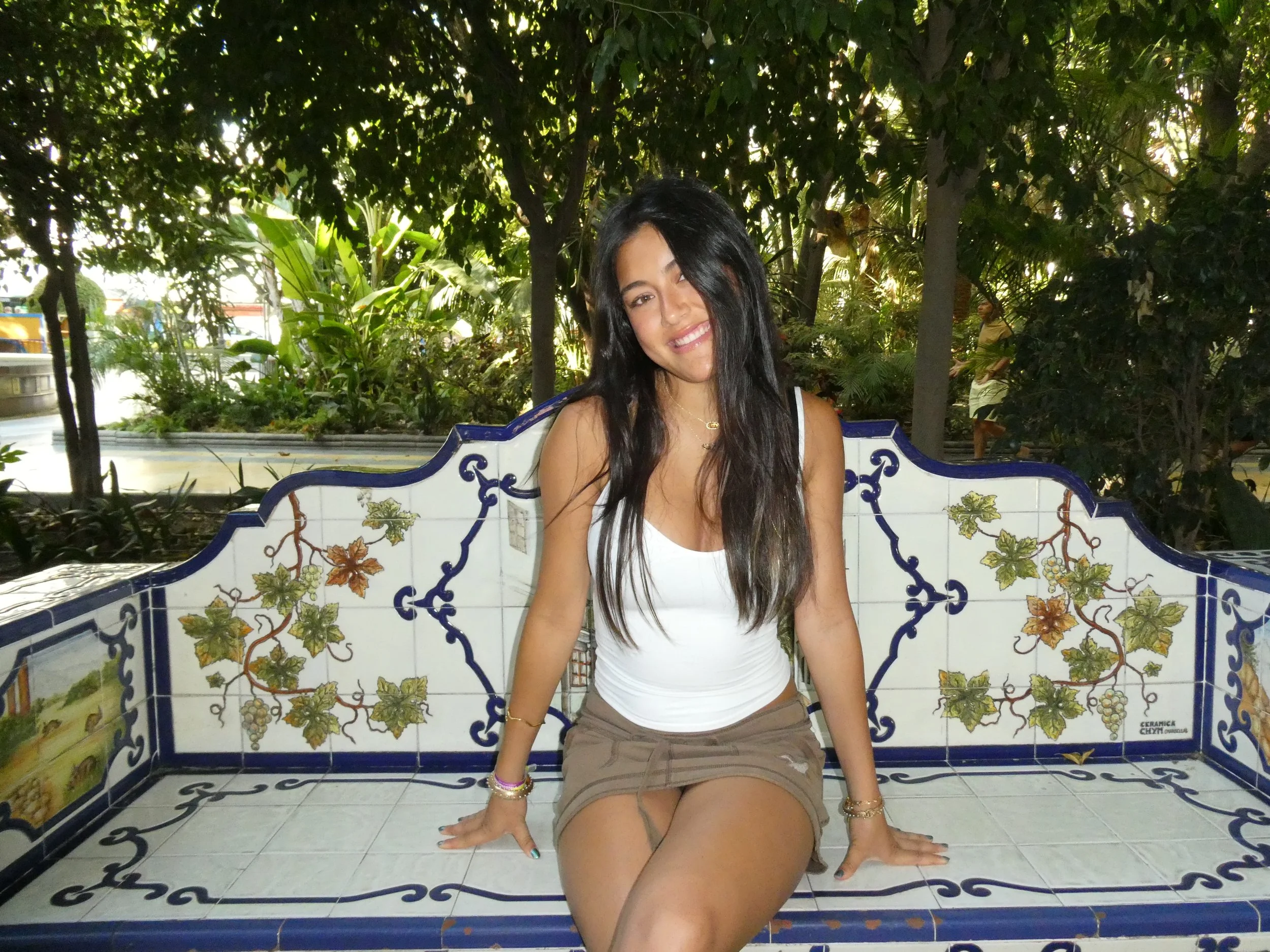 Young woman with long dark hair sitting on a tiled bench outdoors, smiling, surrounded by lush green trees and plants.