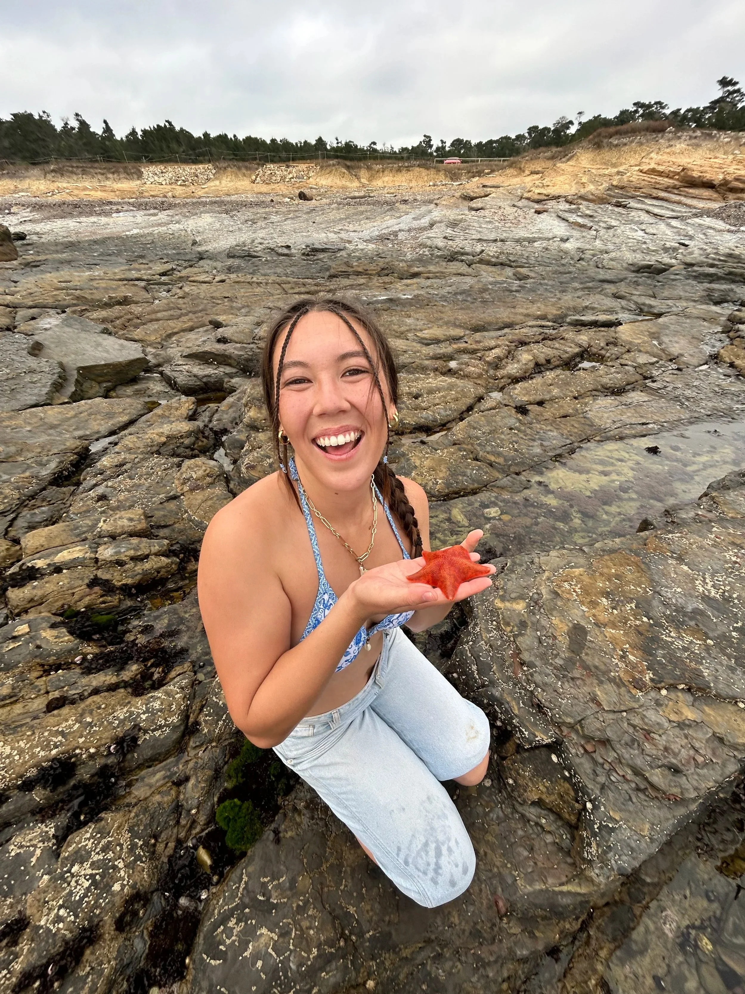 A young woman with braided hair, smiling, wearing a blue swimsuit top and white pants, kneeling on rocky shore, holding a large red starfish, with a cloudy sky and distant trees in background.