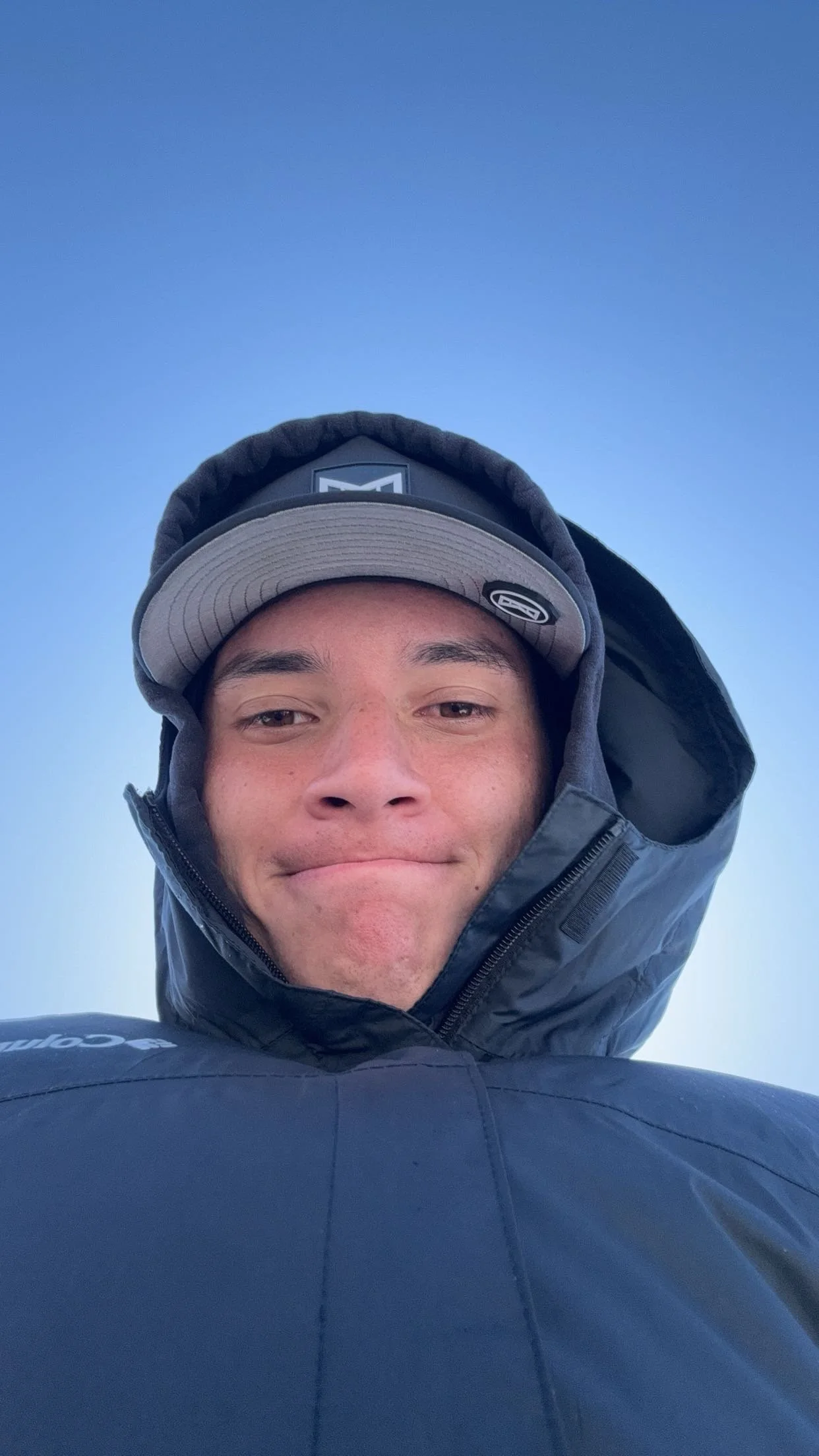 A young man in a black jacket and baseball cap taking a selfie outside against a clear blue sky.