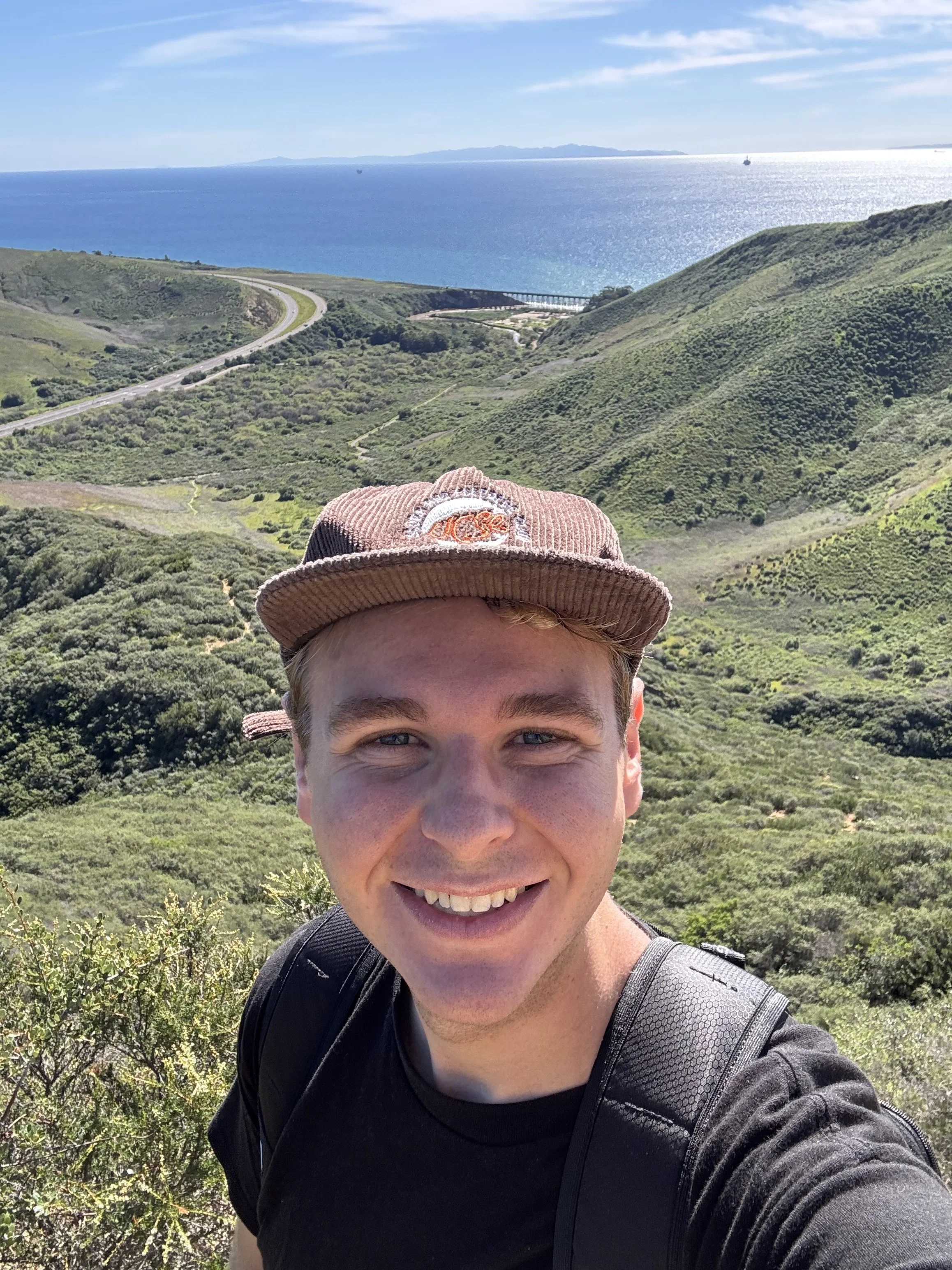 A young man taking a selfie outdoors with lush green hills, a winding road, and the ocean in the background, under a partly cloudy sky.