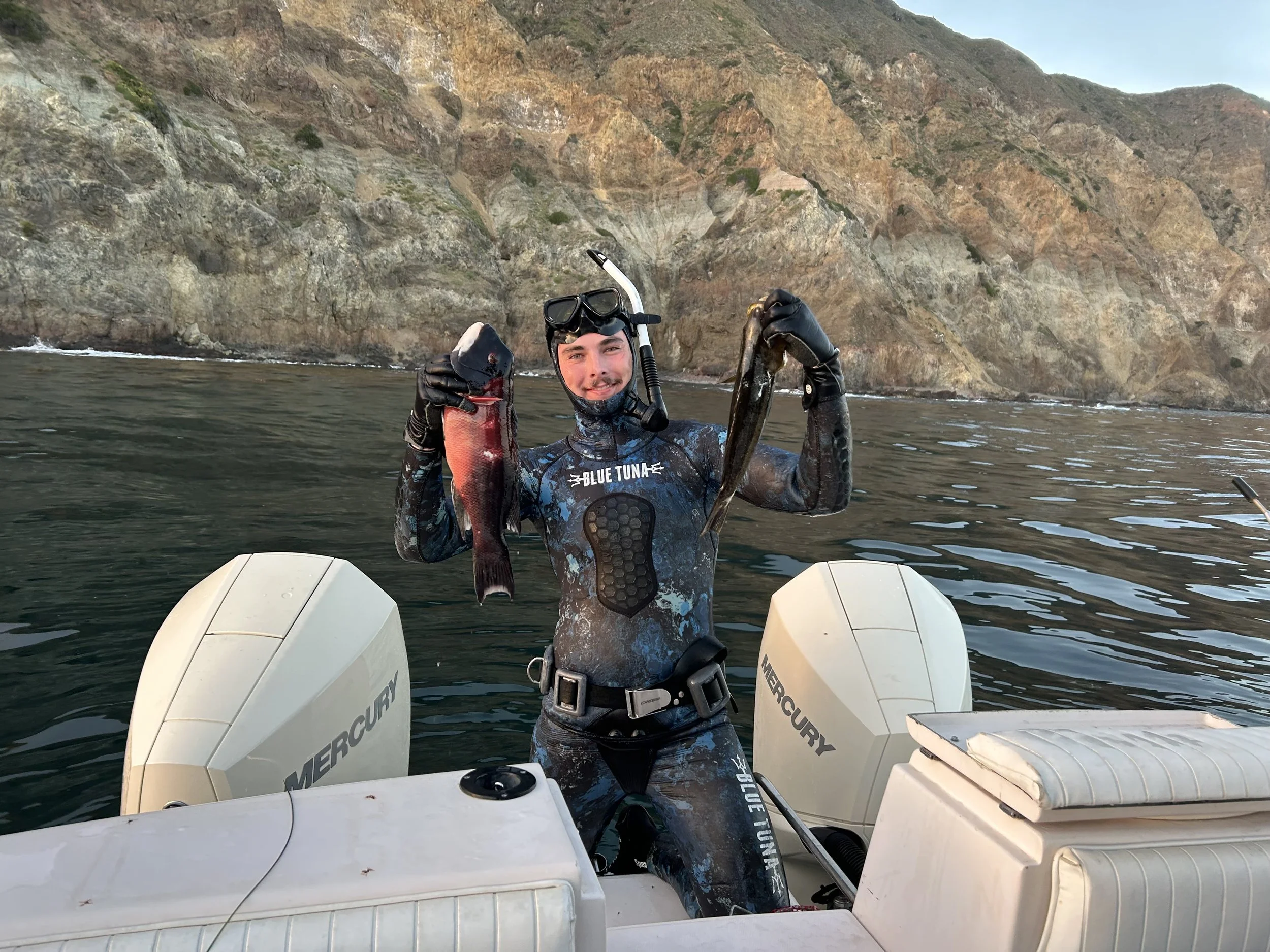 A person in wetsuit holding two fish, standing on a boat in water with rocky cliffs in the background.