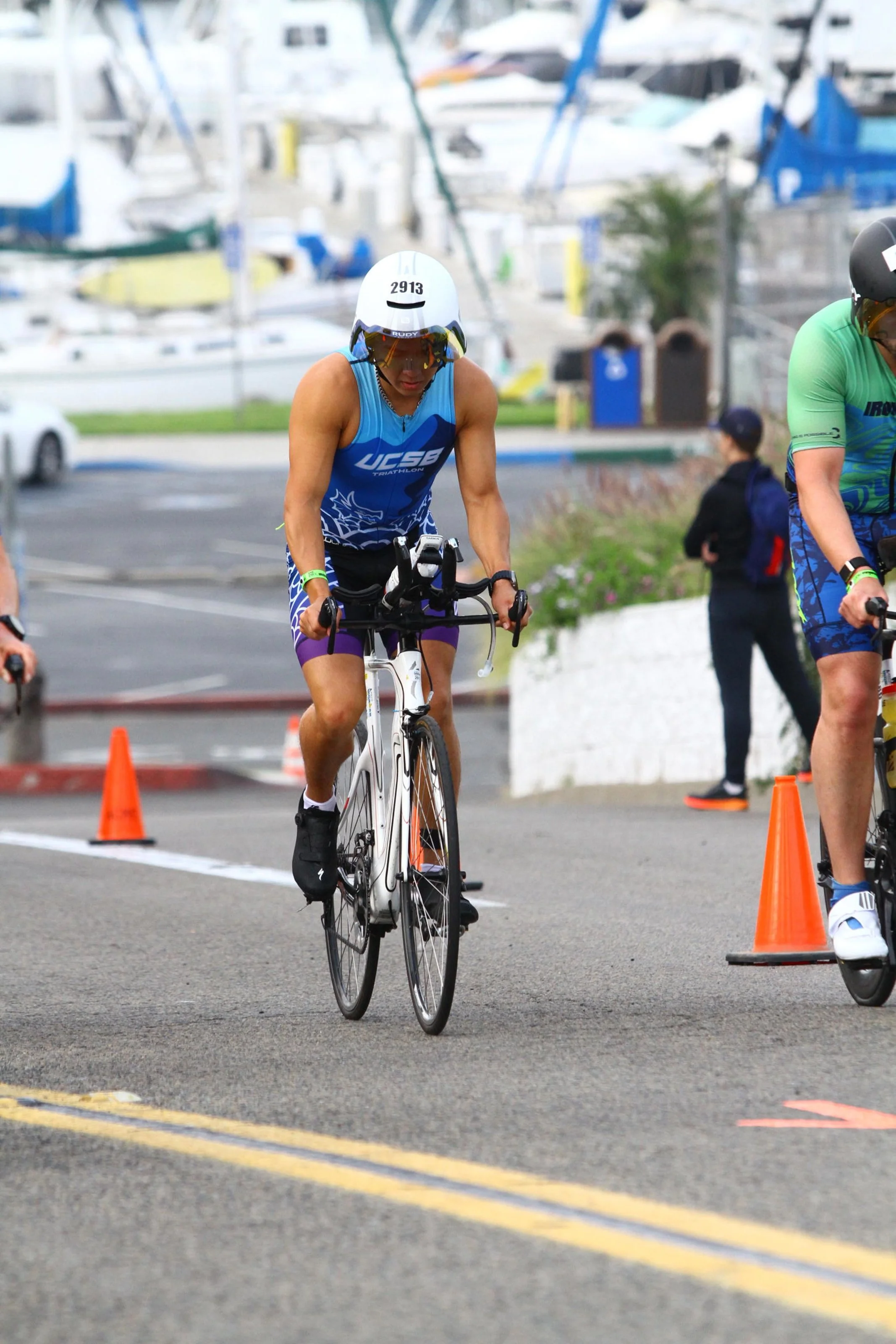 An athlete in a blue triathlon uniform riding a black and white bike during a race, wearing a white helmet and sunglasses, with orange traffic cones and other participants in the background.