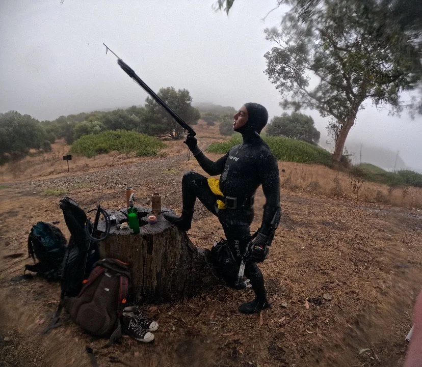 Person in a wetsuit standing on one leg with the other foot on a tree stump, holding a telescoping pole, outdoors in a foggy area with trees and dirt ground, with backpacks, bottles, and other gear on the stump.