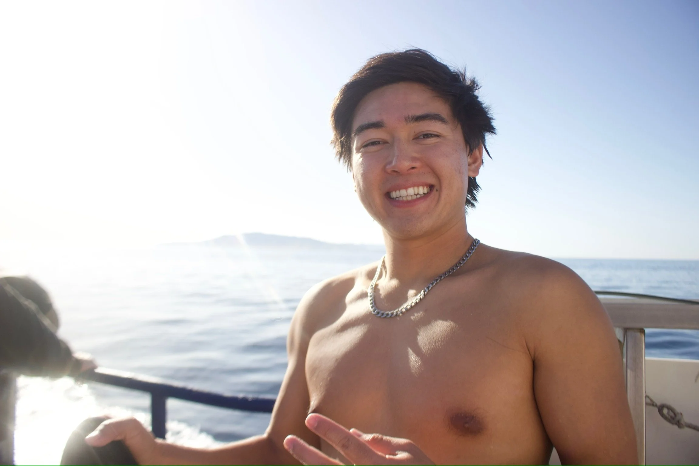 A young man with dark hair and a silver chain necklace smiling on a boat, with open water and a clear sky in the background.