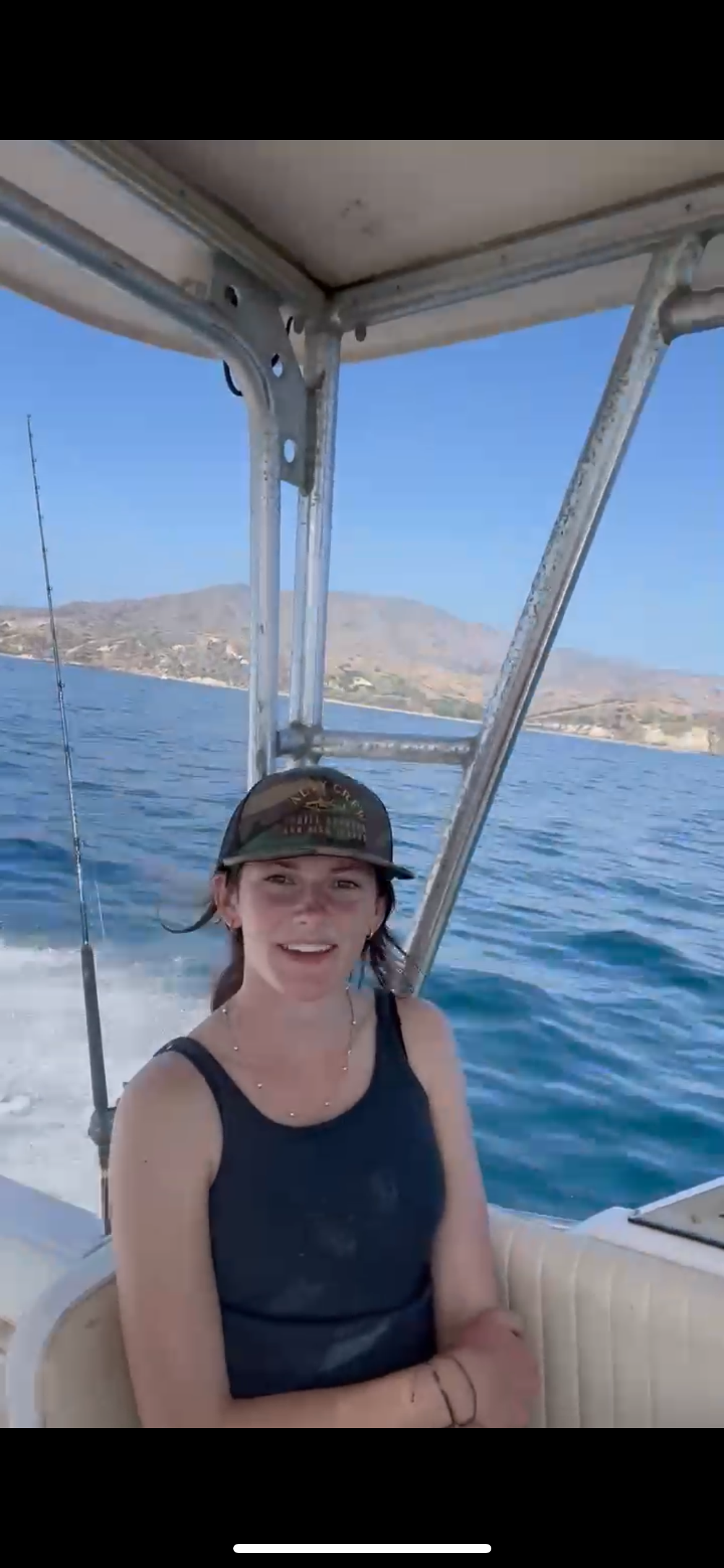 A woman sits on a boat, smiling, with water and distant hills in the background.