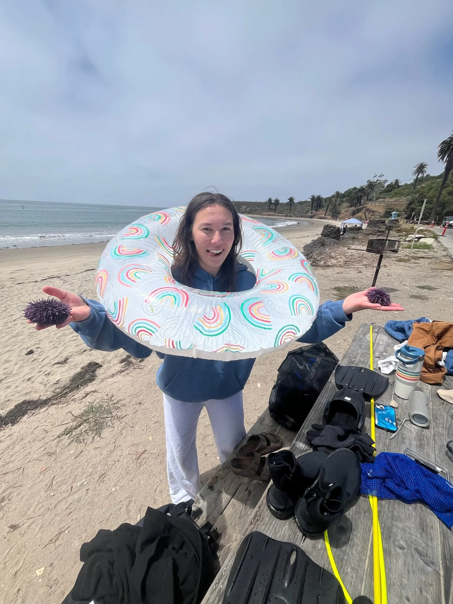 A woman standing on a beach with a colorful inflatable ring around her neck, smiling and holding sea urchins in each hand. There are various items on a picnic table nearby, with palm trees and a cloudy sky in the background.