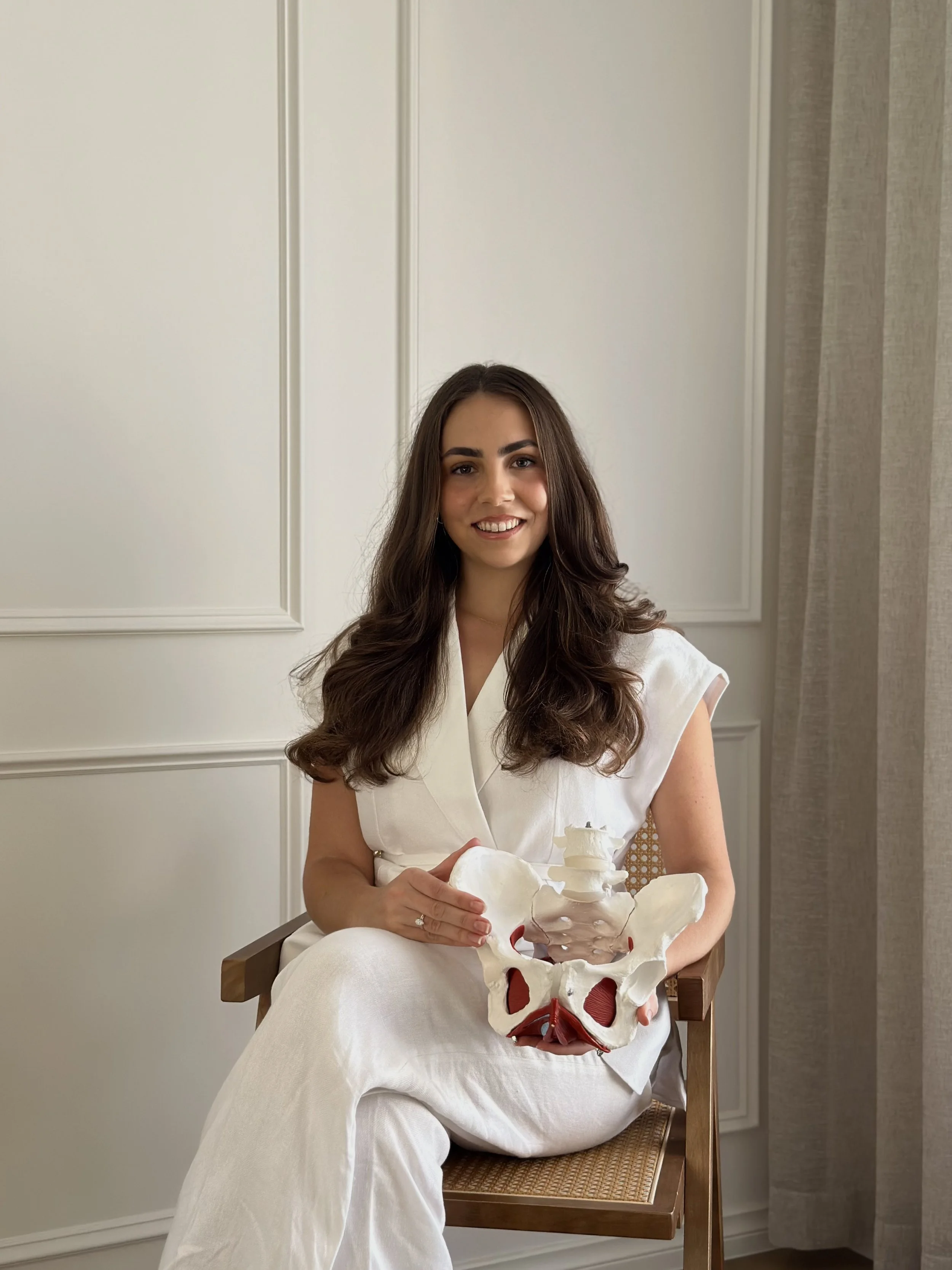 A pelvic floor physiotherapist sitting on a chair, holding a human pelvis model, smiling at the camera, in a room with white paneled walls and a curtain.