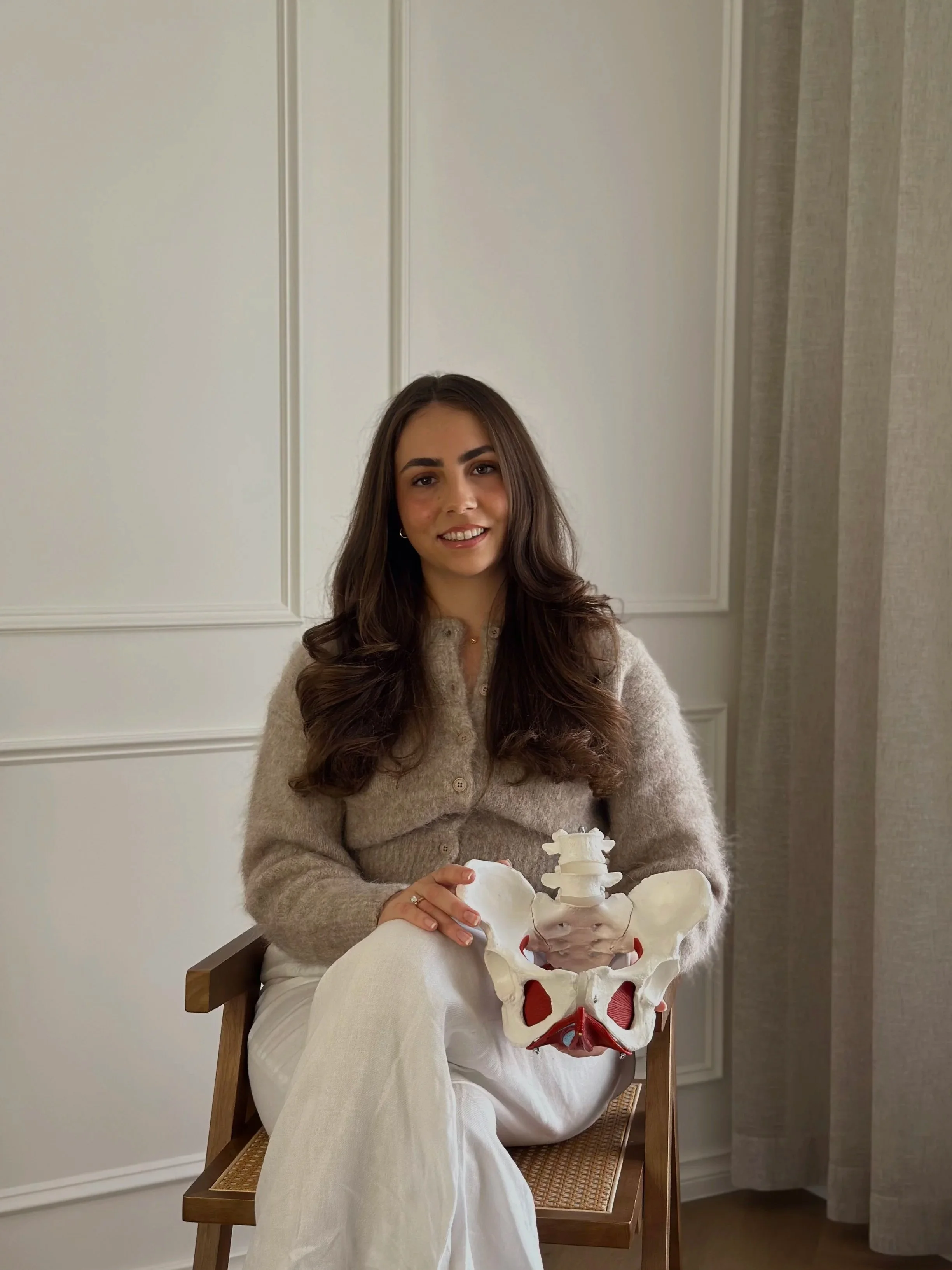 A young woman with long brown hair and a beige sweater sitting on a wooden chair in a room, holding a replica of a human pelvis.