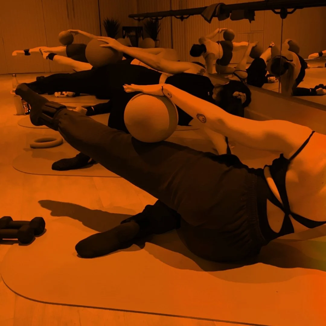 People participating in a yoga or fitness class, lying on mats, with desktop ball equipment, in a dimly lit room with mirrors.