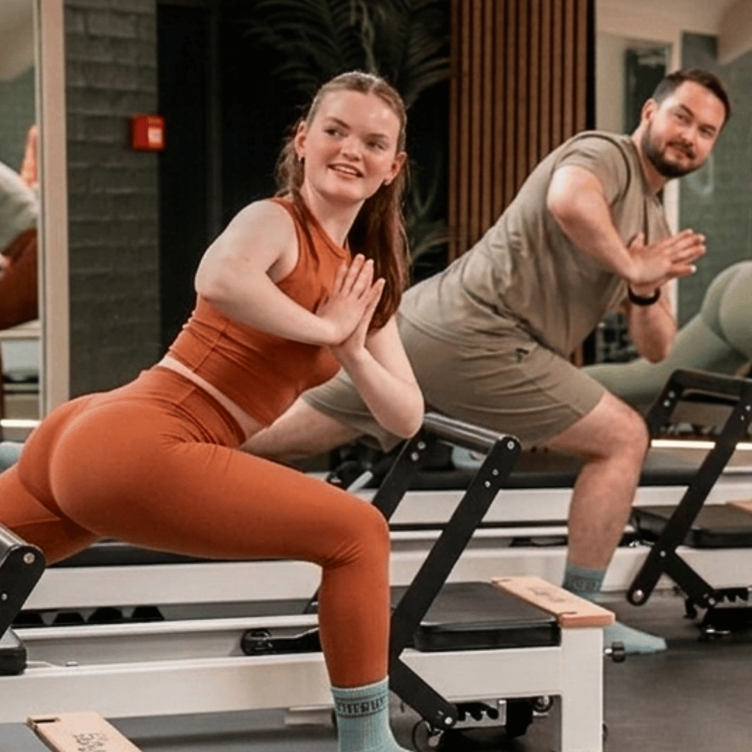 Four women in white workout clothes laughing and smiling together in a Reformer Pilates studio.