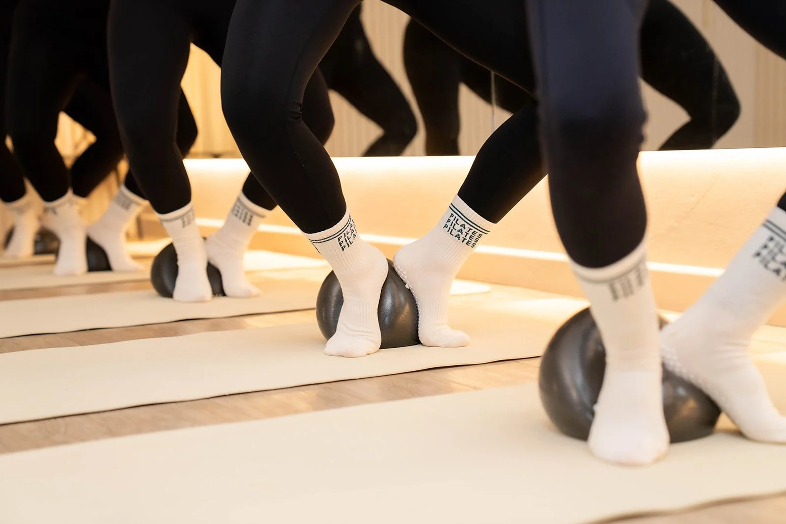 People participating in a Pilates class using small black exercise balls, wearing matching white socks and black leggings, on white exercise mats in a studio.
