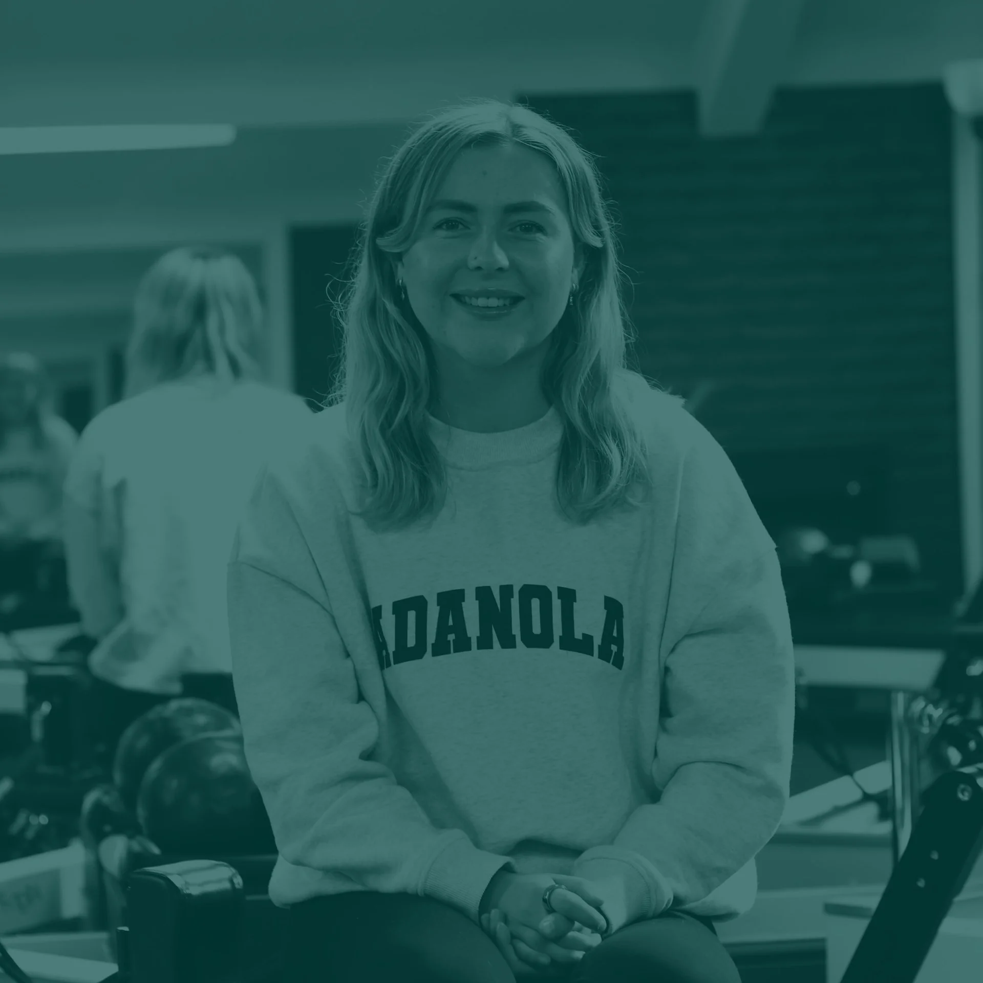 A smiling woman with blonde hair wearing a sweatshirt that says 'Madana' sitting in a gym or fitness center with weights and exercise equipment in the background.