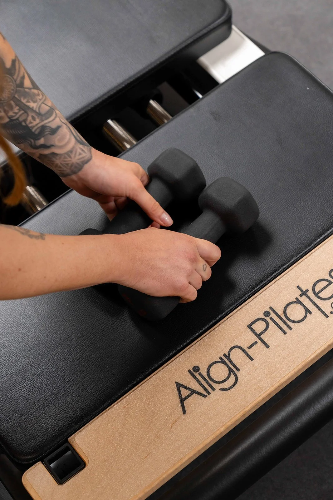 Person with tattoos lifting a black dumbbell in a gym. The dumbbell is on a black workout bench, with a wooden sign reading 'Aligned-Plate' nearby.