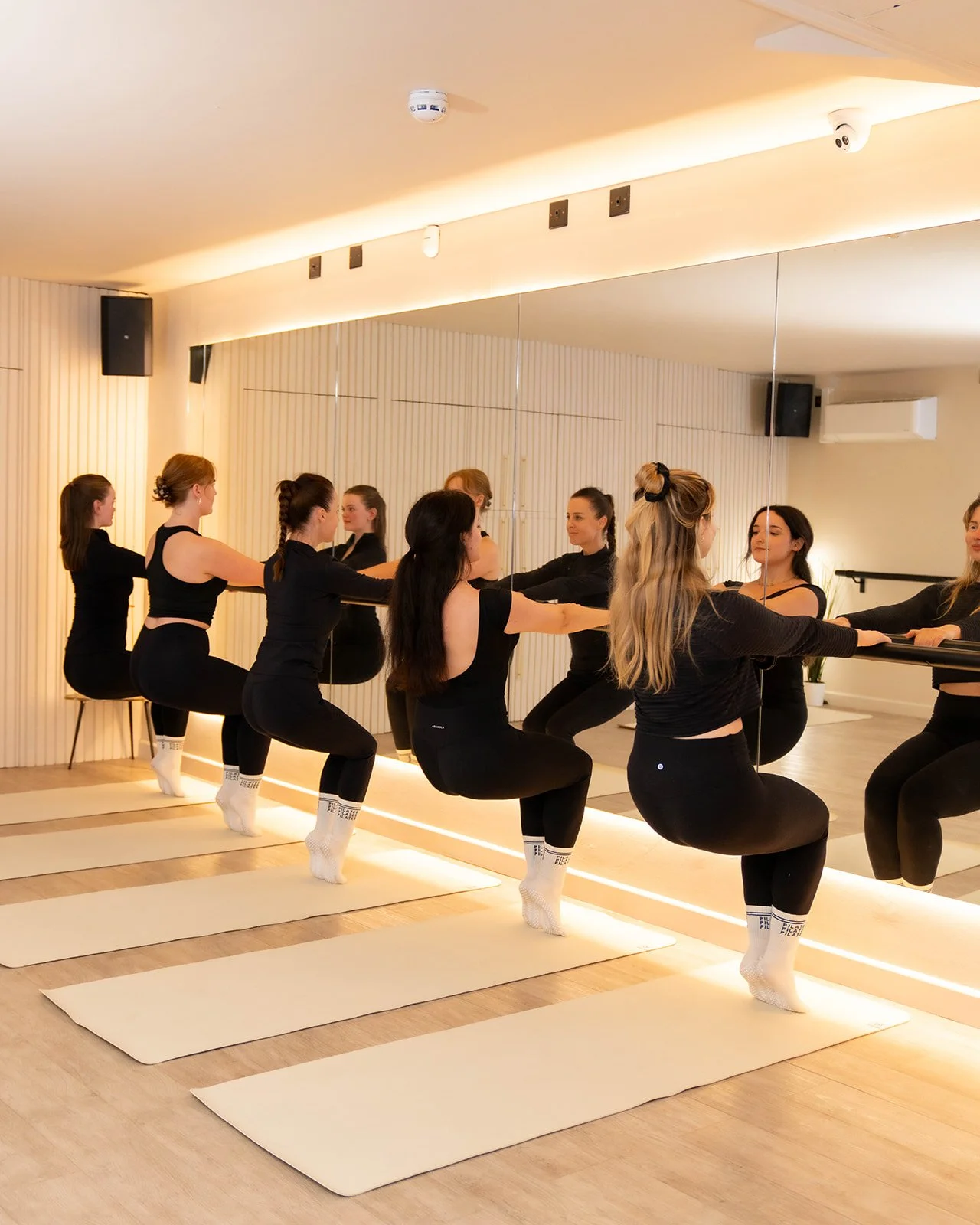 A group of women dressed in black workout clothes are doing a synchronized exercise in a mirrored barre fitness studio. They are kneeling on mats with their arms extended forward, holding onto a barre or similar equipment.