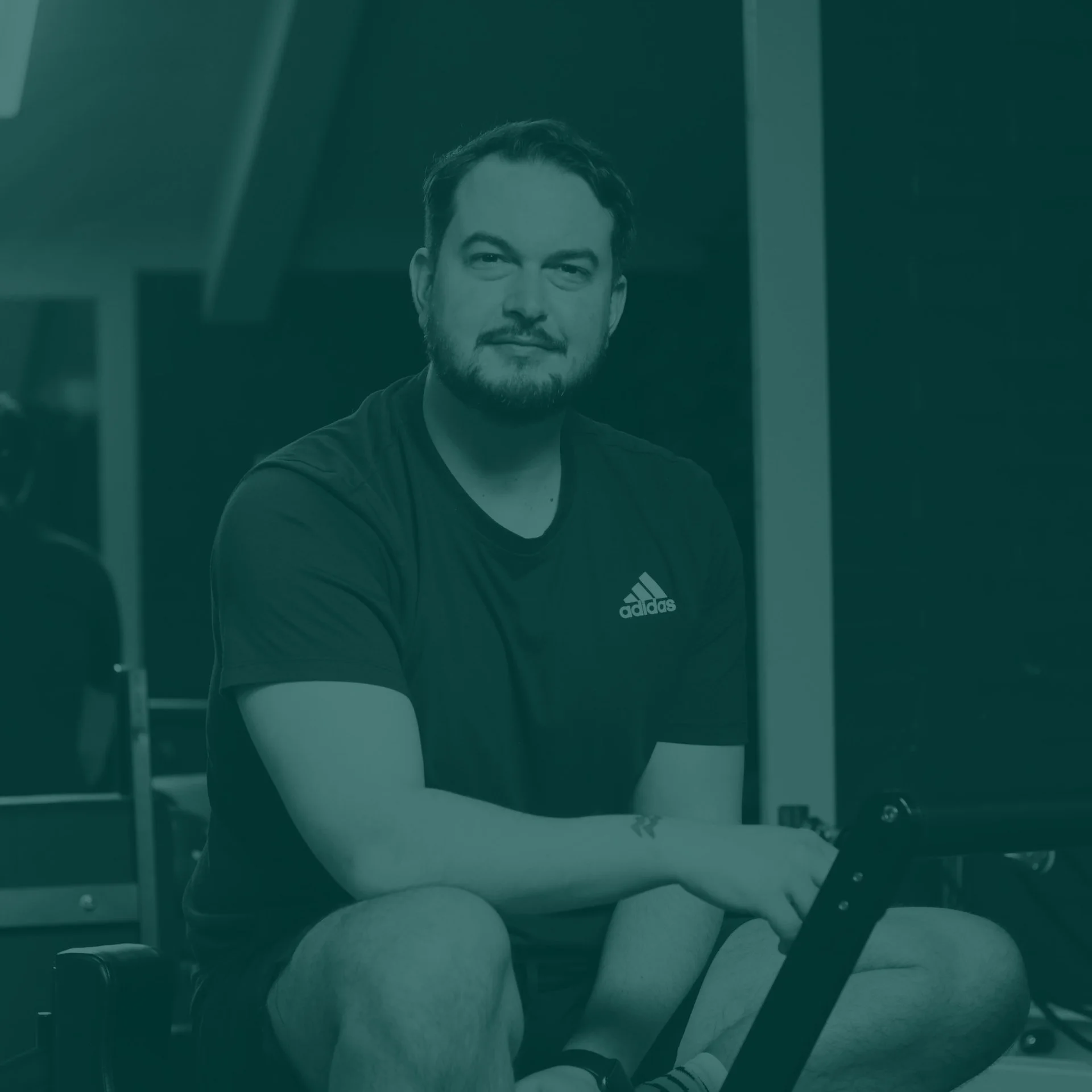 A young man with a beard sitting cross-legged in a gym or fitness area, wearing a black Adidas t-shirt, looking at the camera with a slight smile, with gym equipment in the background.
