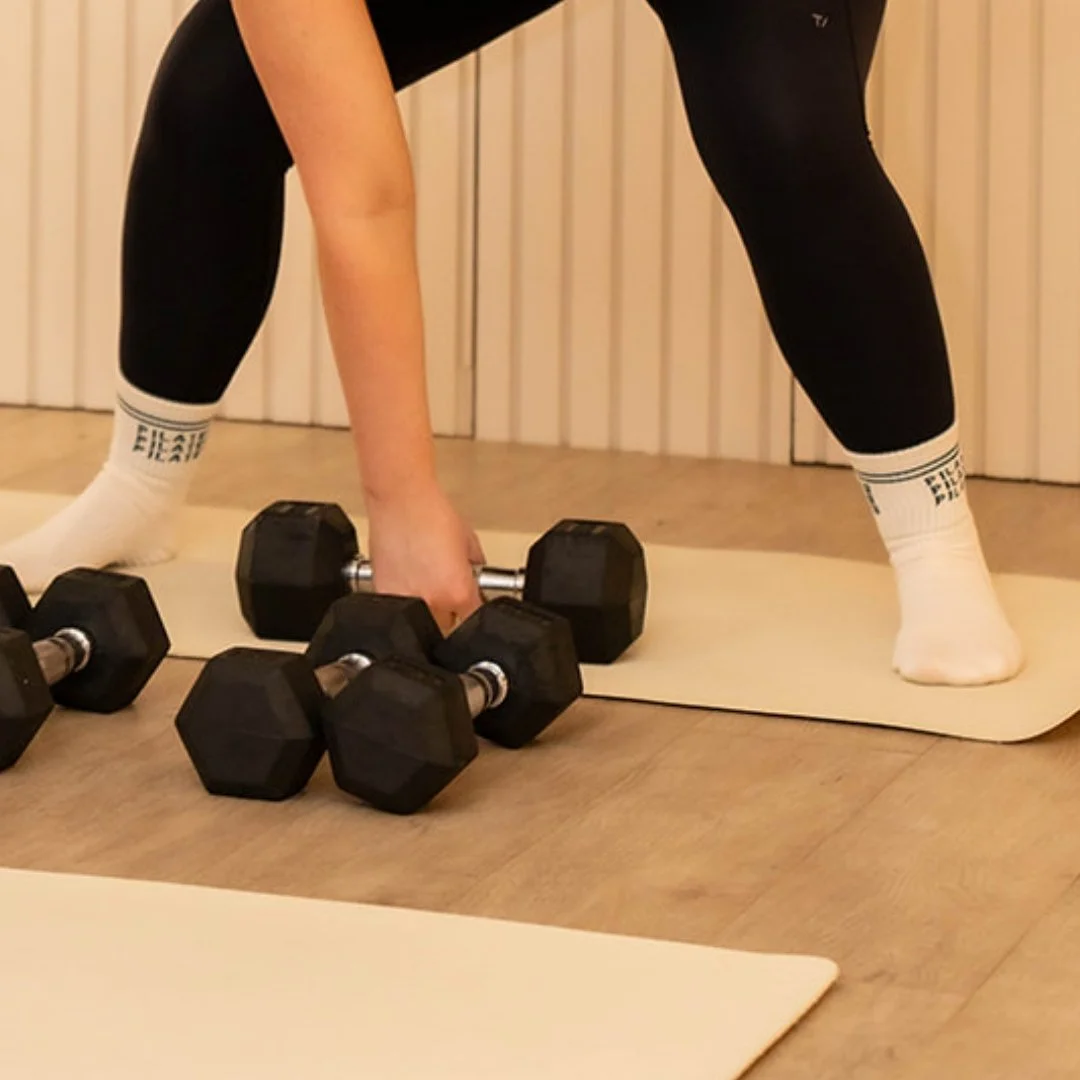 Person in workout clothes lifting a dumbbell in a bent-over position with additional dumbbells on the floor nearby on a yoga mat.