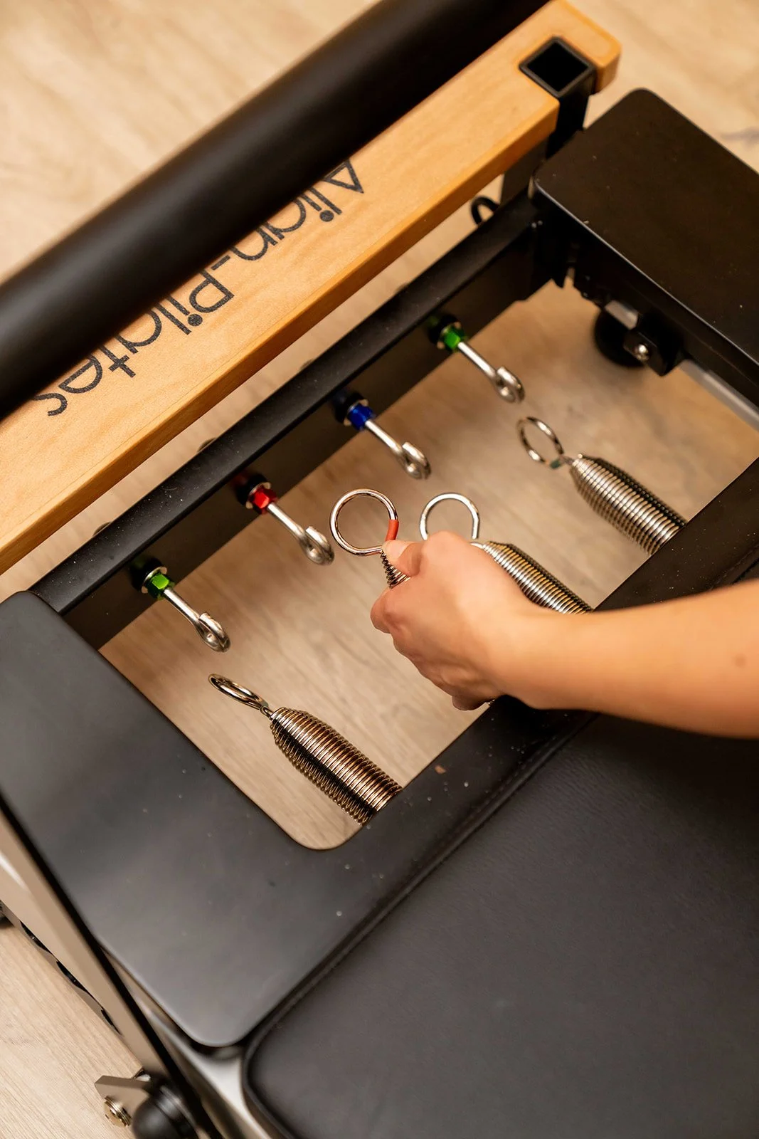 Close-up of a person's hand adjusting a metal spring on a foot pedal machine, with foot pedals and springs visible, on a wooden floor.