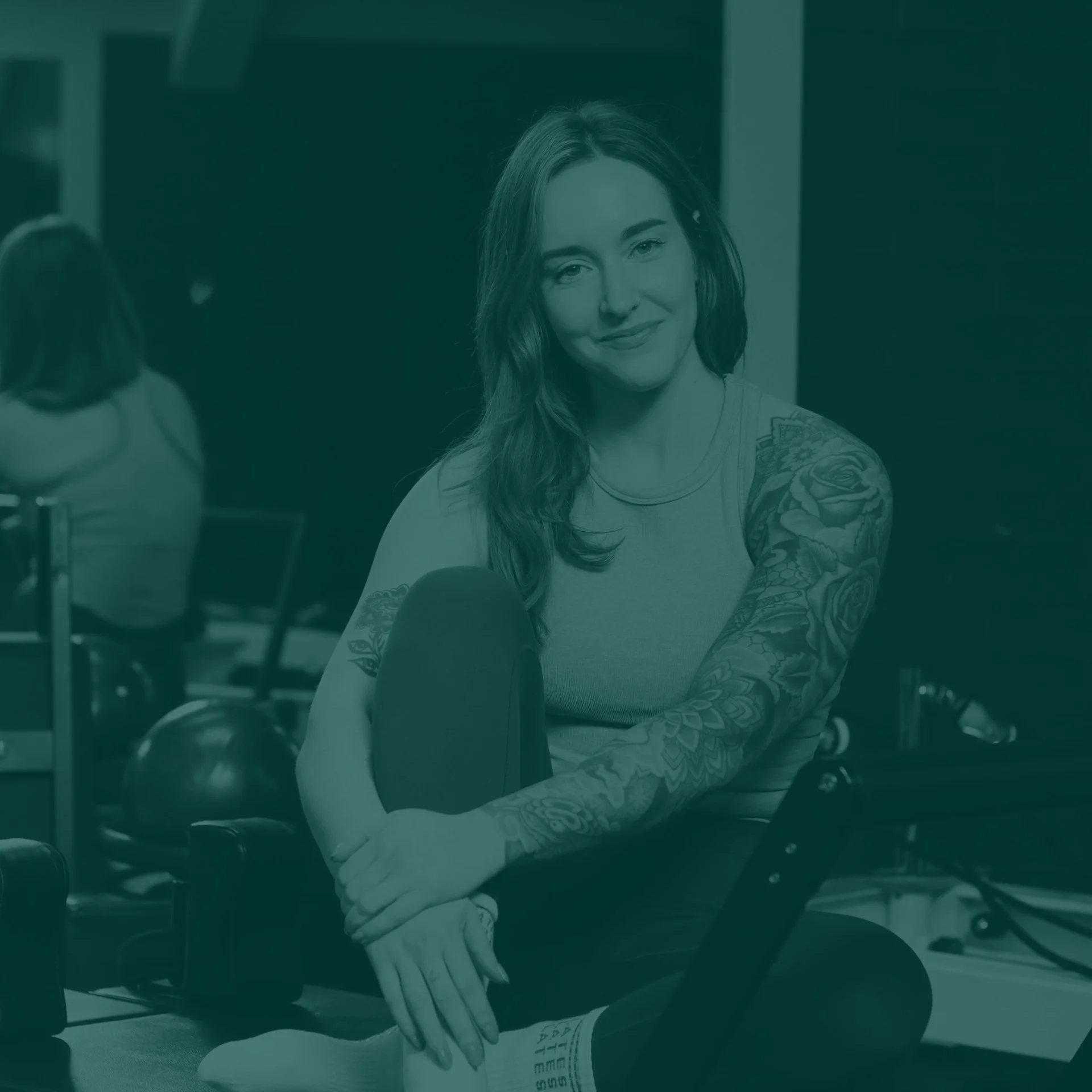 A woman with long hair and tattoos sitting in a gym, smiling at the camera.