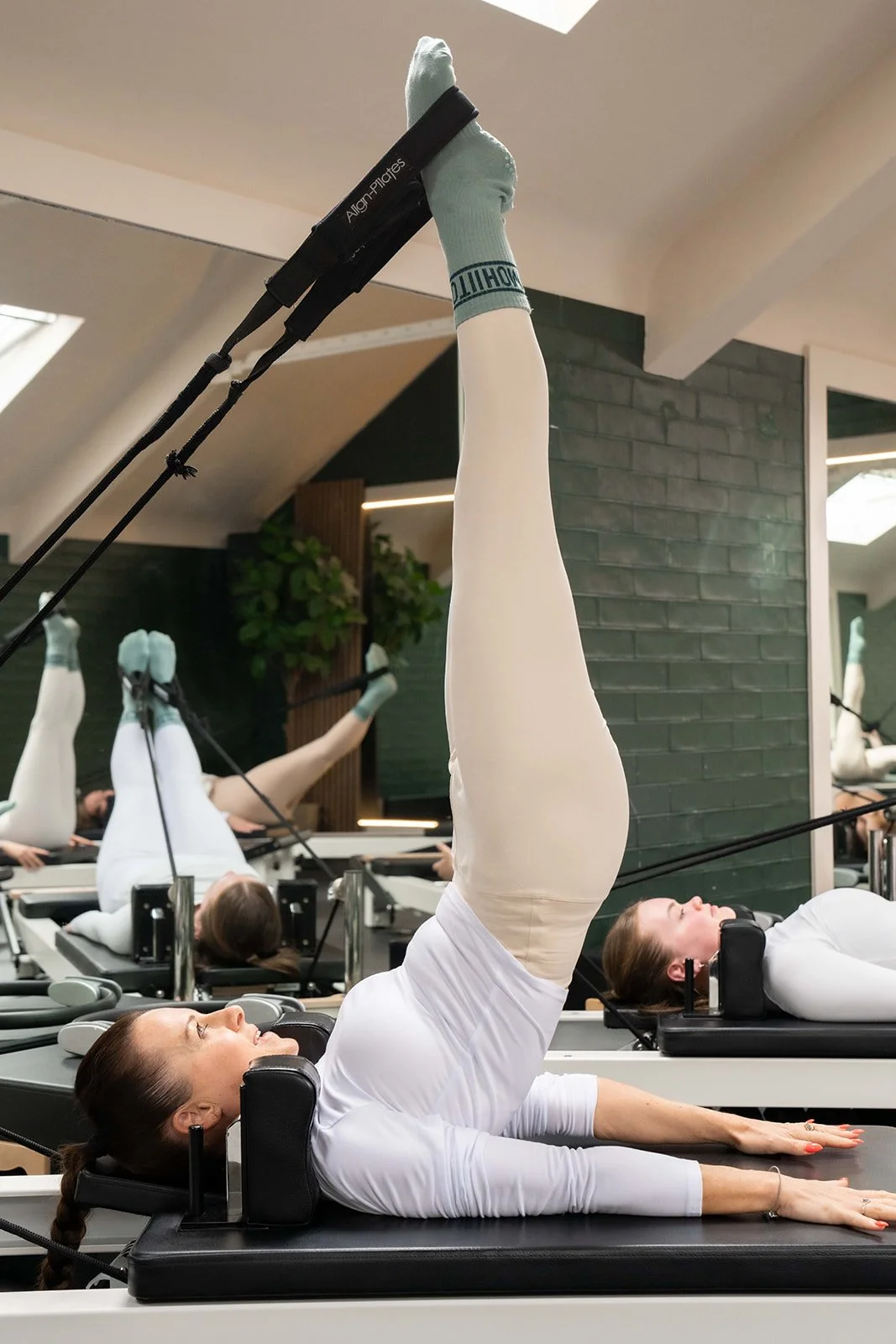People exercising on Pilates reformer machines in a fitness studio, with their legs elevated and using resistance straps.