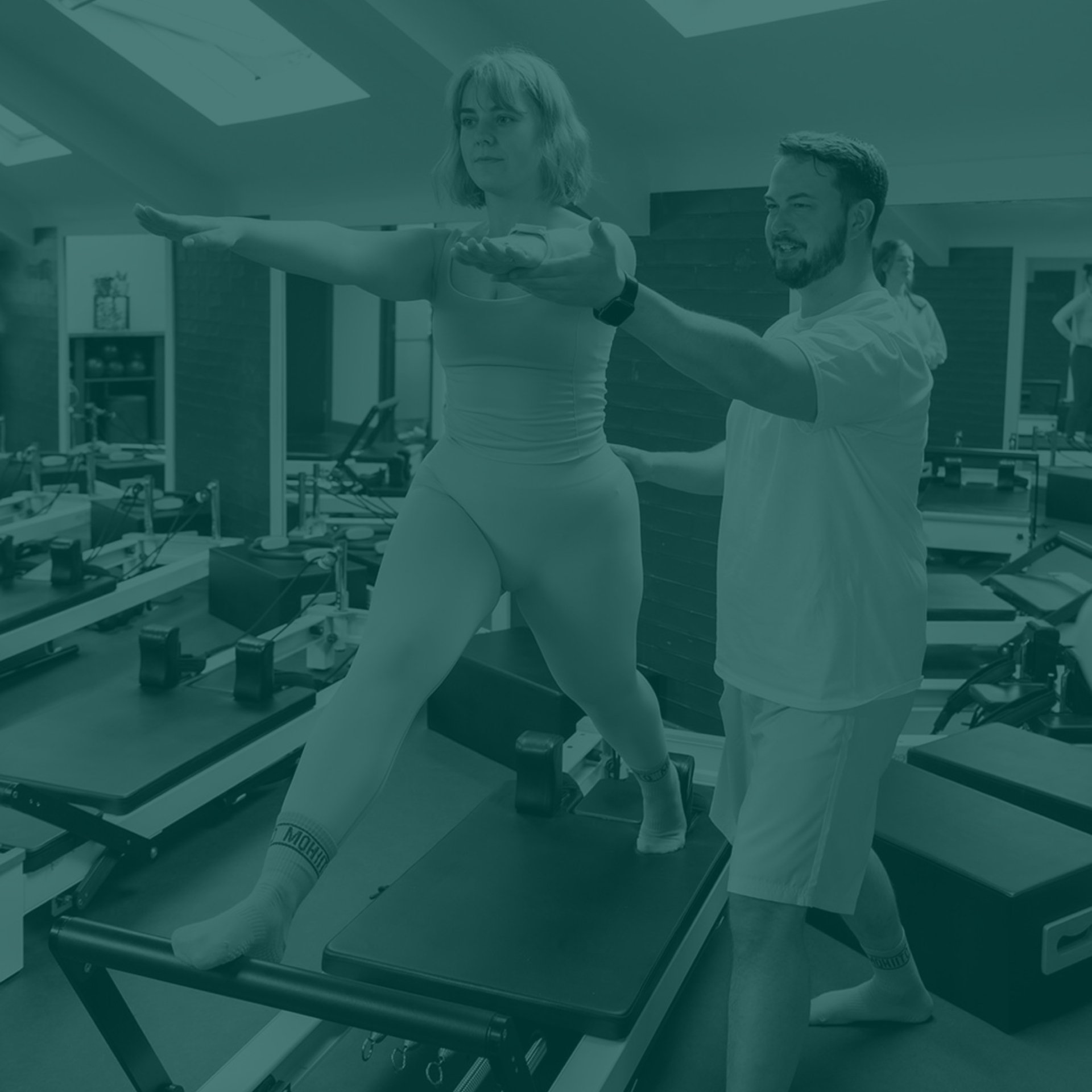 A woman performing a balance exercise on a Pilates reformer machine while a man assists her in a fitness studio.