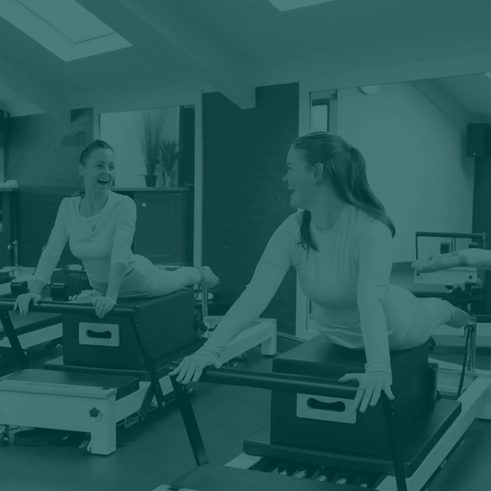 Two women smiling and doing Pilates exercises on reformer machines in a fitness studio.