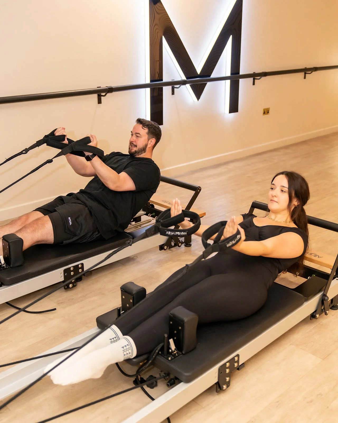 A man and a woman using Pilates reformer machines in a fitness studio.
