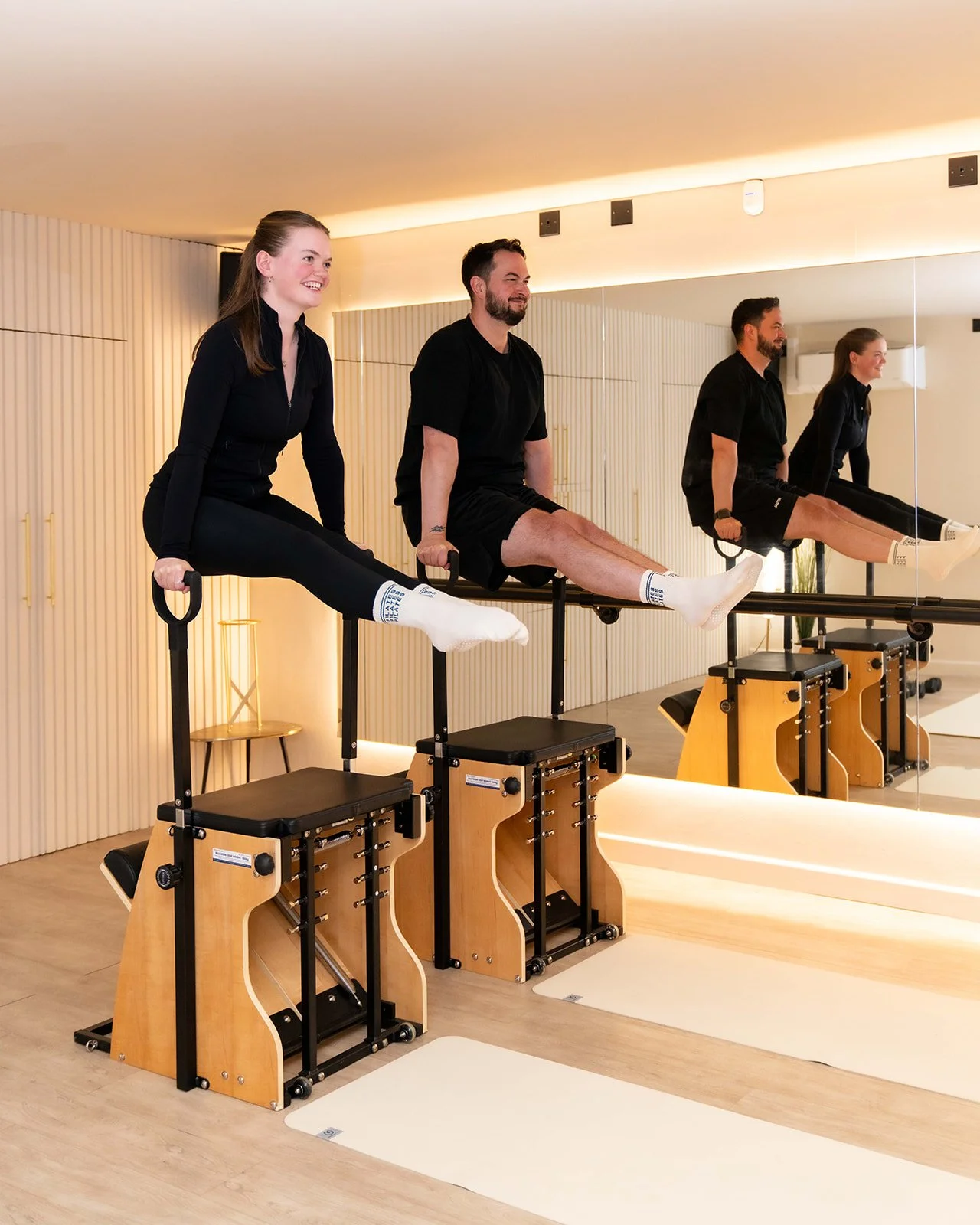 Three people in black workout clothes, two women and one man, doing Pilates on reformer machines in a fitness studio with wood paneling and a large mirror.