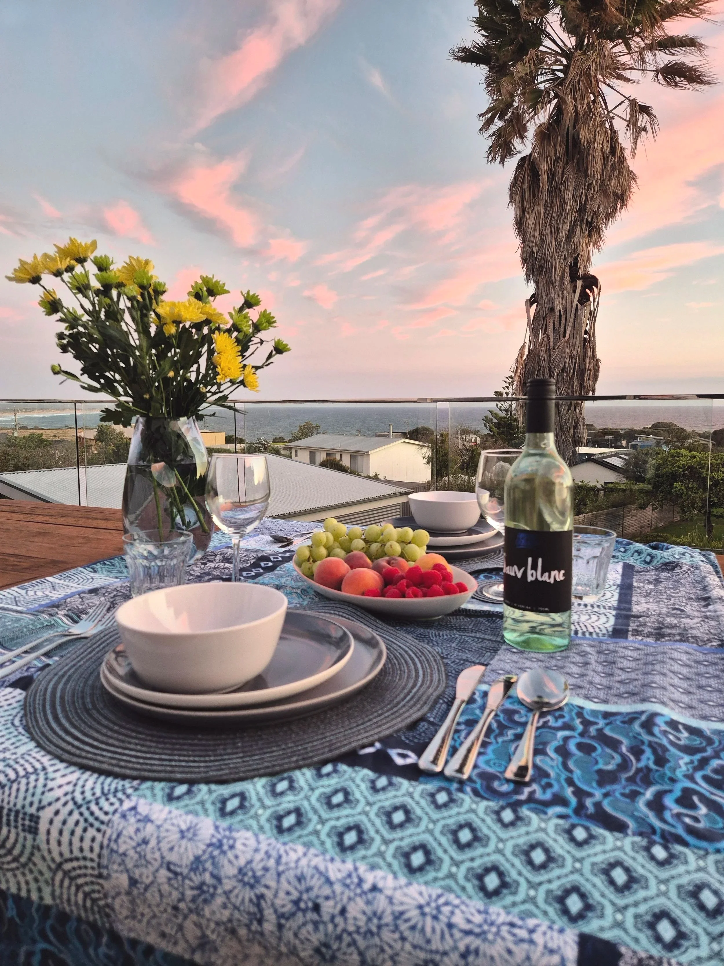 A table set with plates, bowls, glasses, and utensils, with a decorative tablecloth, a bowl of fresh fruit, a vase of yellow flowers, and a bottle of Georgie Orbach Pinot Gris, overlooking a scenic view of houses, trees, and the ocean during sunset.