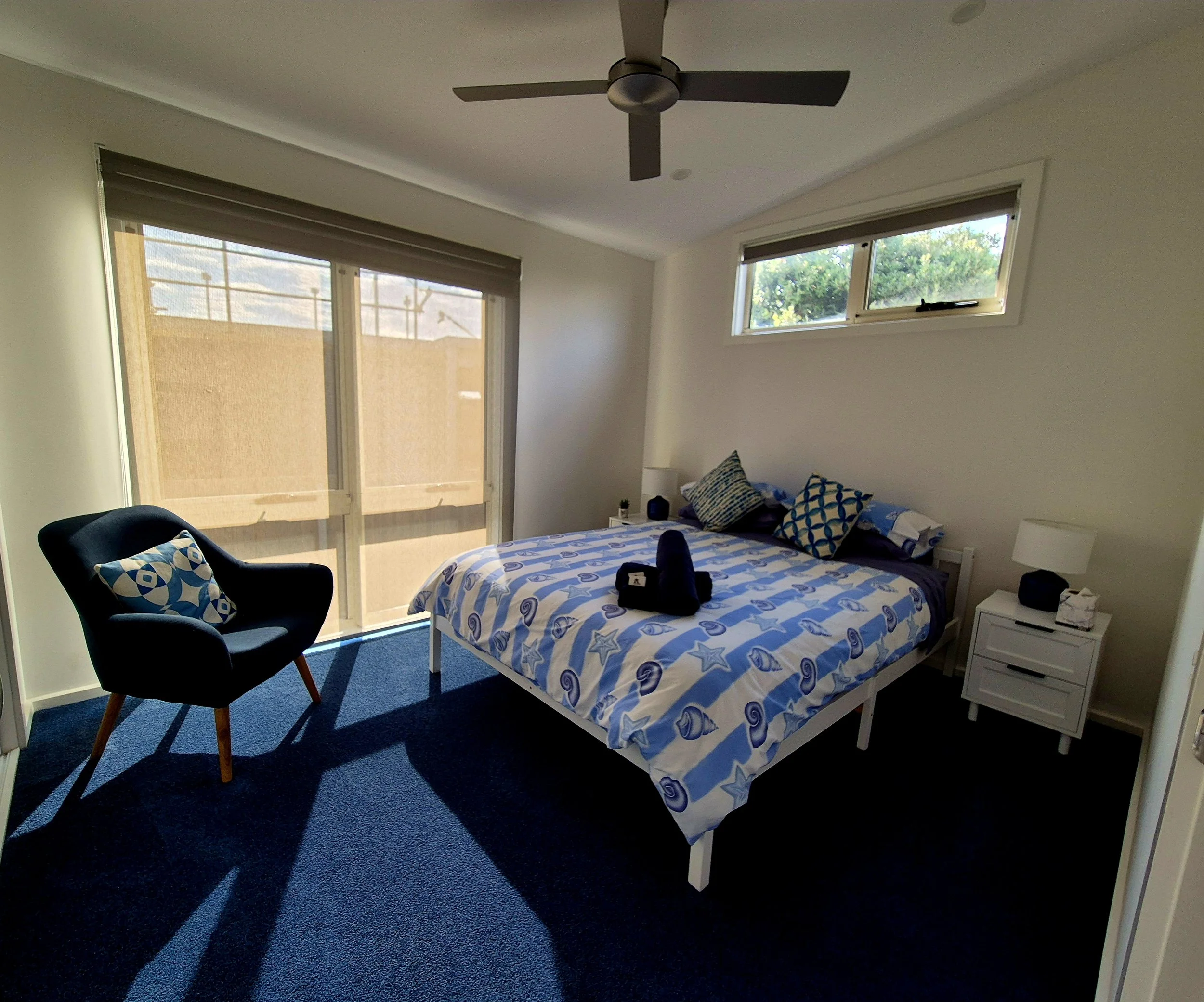 A bedroom with a bed, armchair, and windows. The bed has blue and white patterned bedding with matching pillows. The armchair is dark with a patterned cushion. The windows have blinds and let in some natural light. There is a small bedside table with a lamp and a few items.