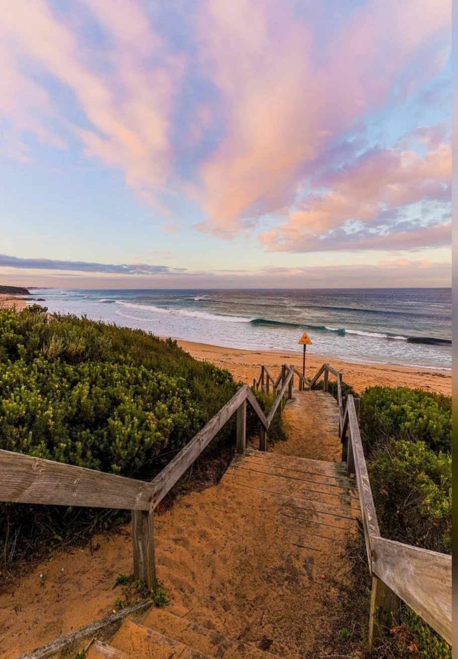 A wooden walkway with steps covered in sand leading down to the beach at sunset