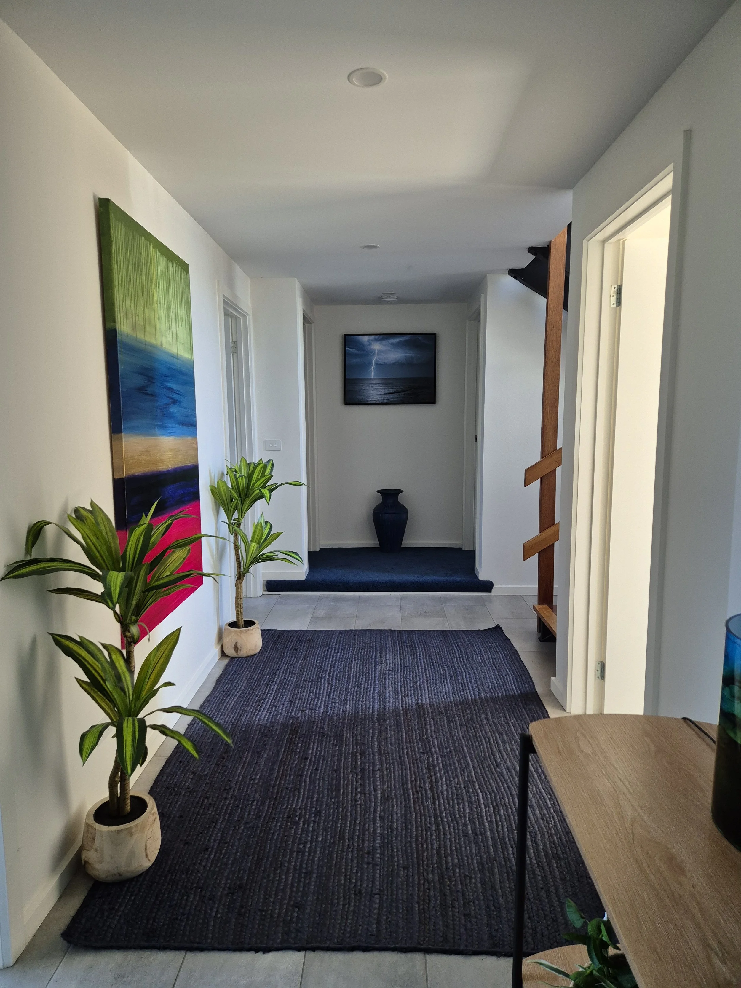A hallway with a dark woven rug, potted plants, colorful artwork, a blue vase, a framed lightning storm photo on the wall, and a small wooden table.
