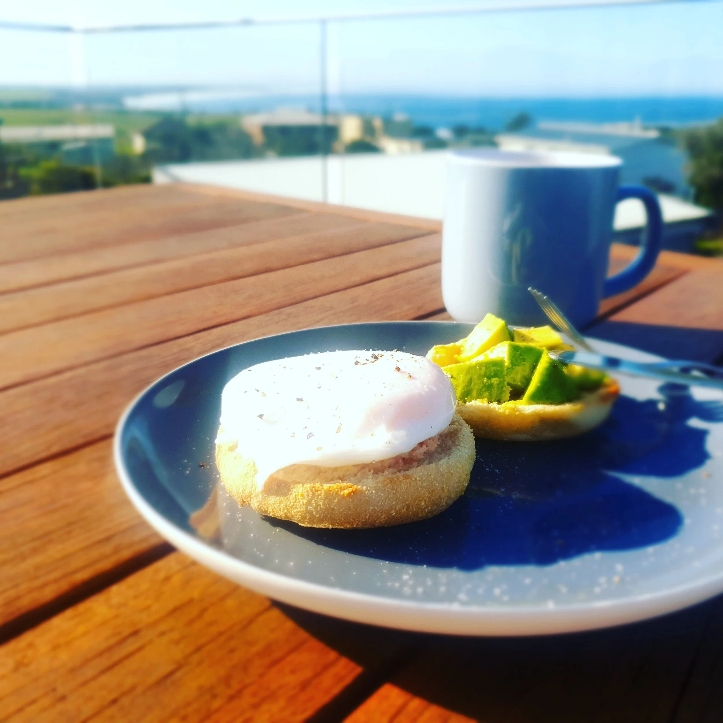 Poached egg and avocado on English Muffins and a coffee on the table on the deck, looking toward the ocean views in the background.