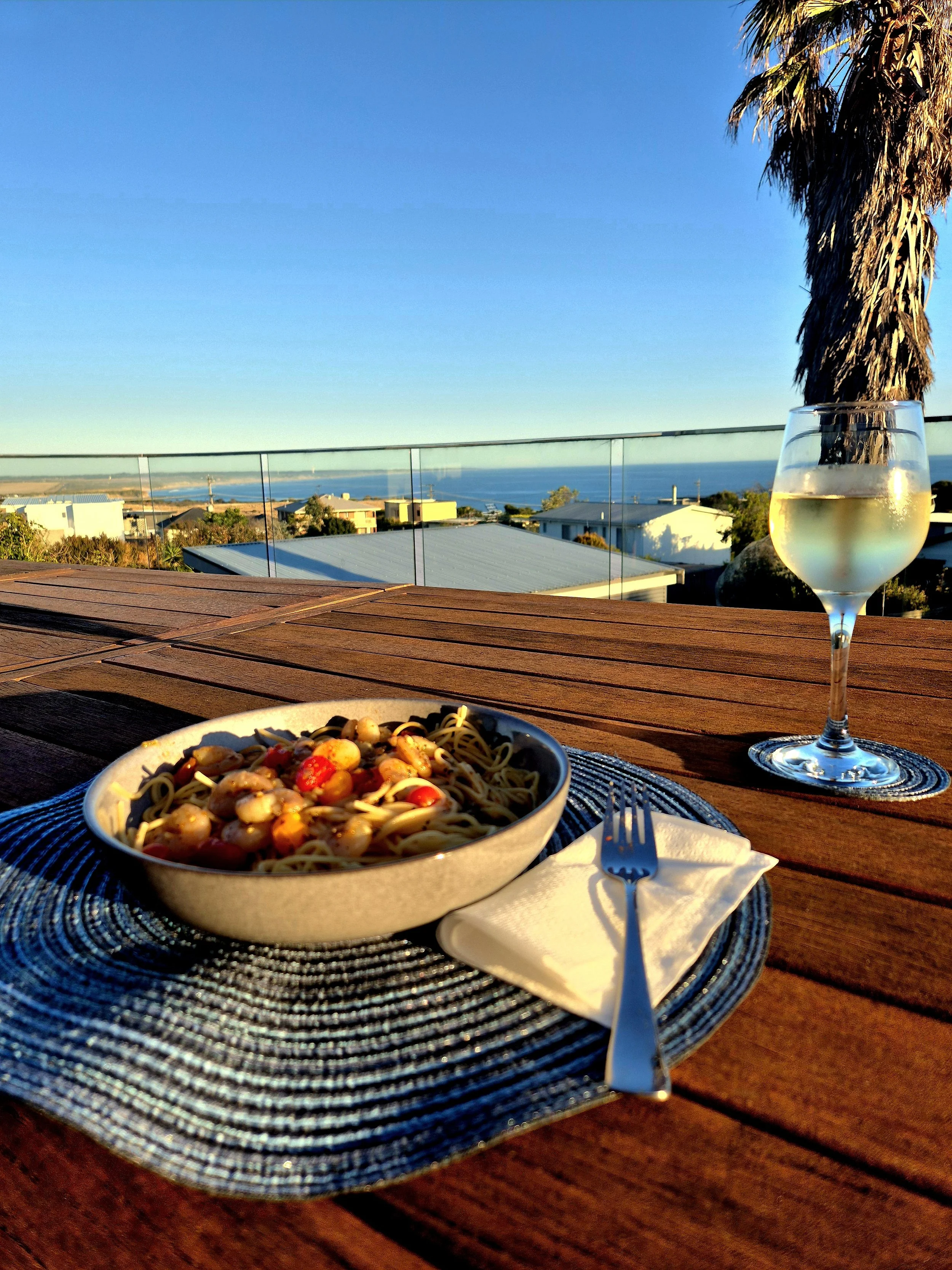 Picture of the wooden table on the deck at Vista Azure featuring a bowl of pasta, glass of wine and beautiful seaside views.