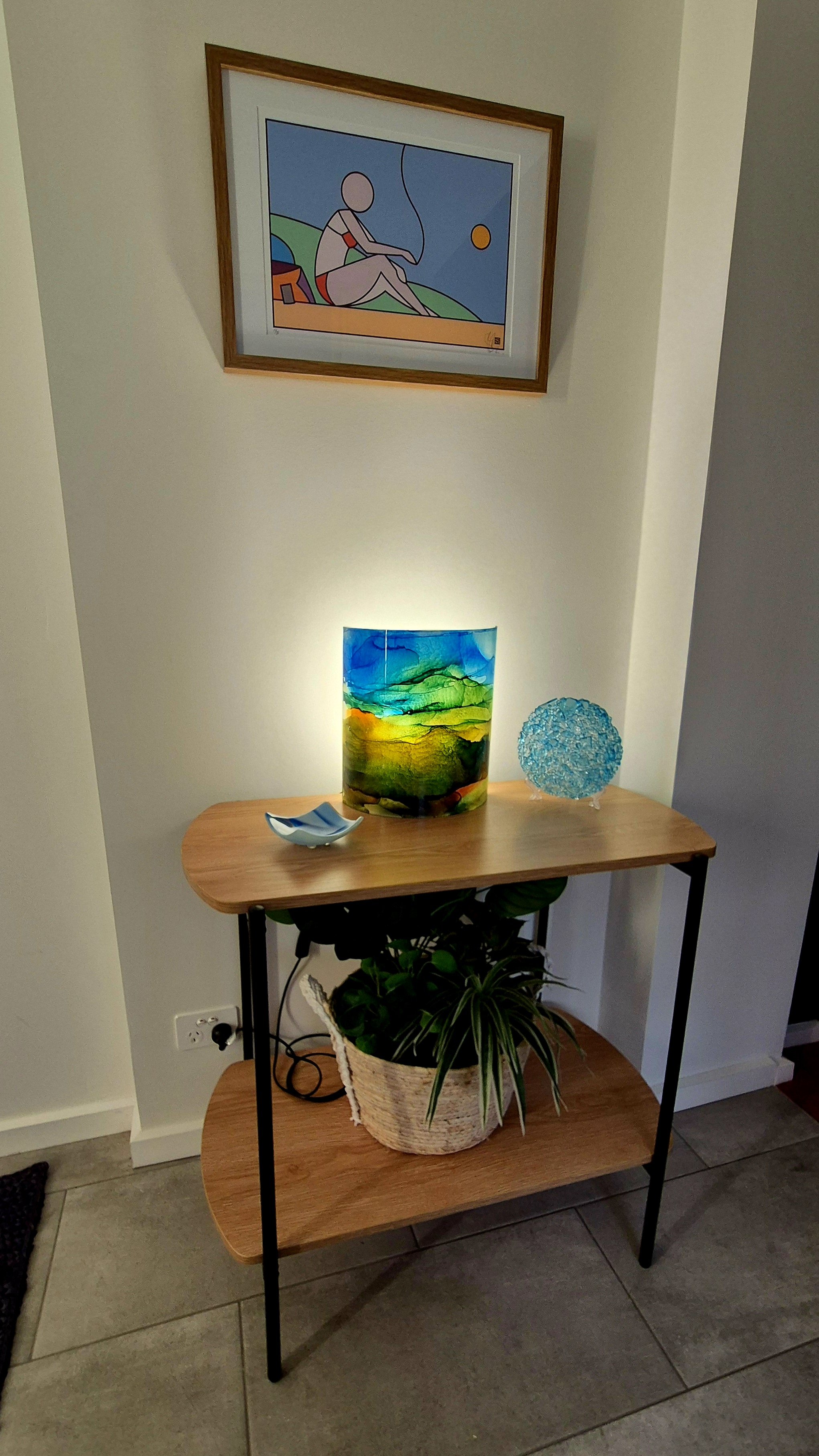 A wooden and metal shelf unit with two levels. On the top level, a colorful glass lamp, a small decorative bowl, and a blue textured sphere. On the bottom level, a potted plant. A framed abstract painting hangs on the wall above the shelf, and the se