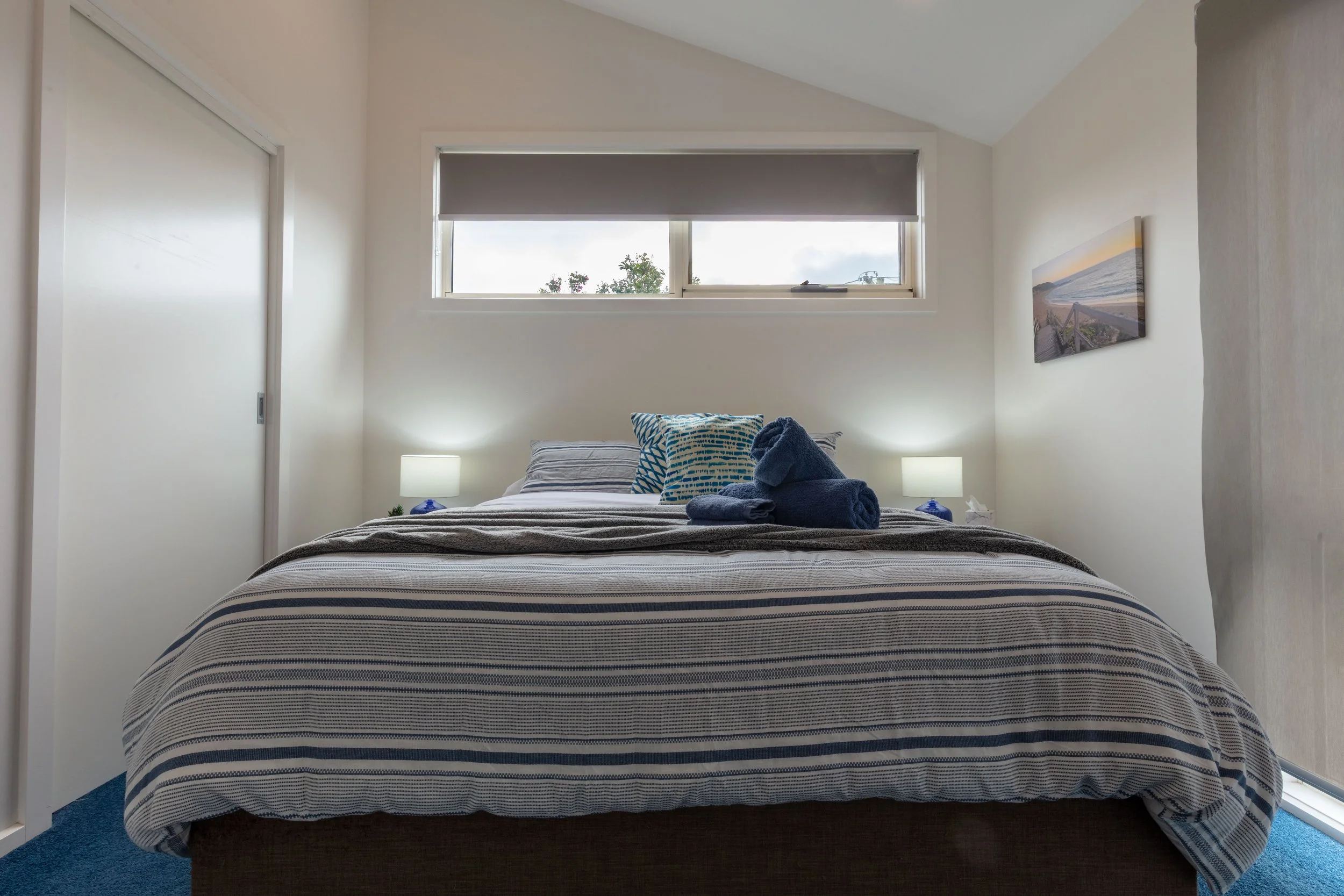 A bedroom with a bed, armchair, and windows. The bed has blue and white patterned bedding with matching pillows. The armchair is dark with a patterned cushion. The windows have blinds and let in some natural light. There is a small bedside table with a lamp and a few items.