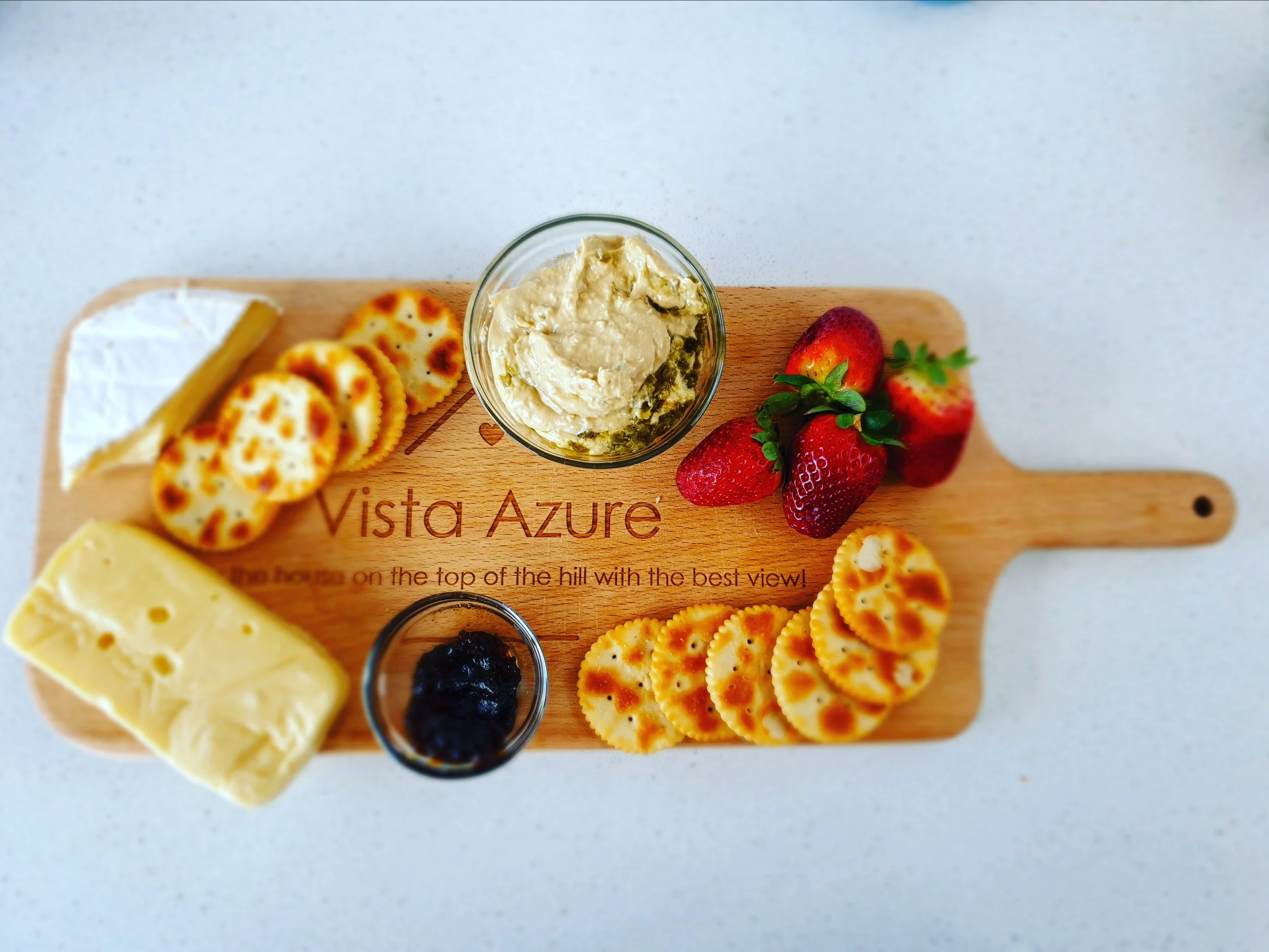 A cheese board featuring the name of the holiday home Vista Azure, decorated with cheese, crackers, hummus and strawberries. 