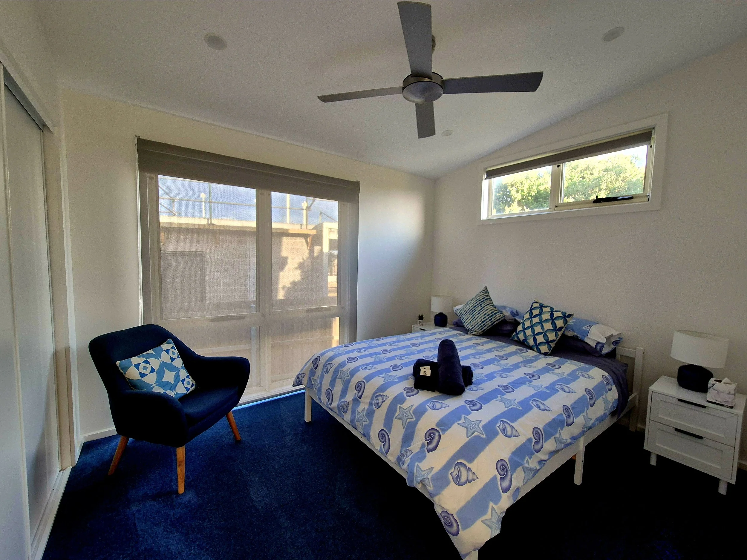 A bedroom with a bed featuring blue starfish and swirl patterns on the bedding, a black armchair with a matching blue and white geometric-patterned pillow, a small white nightstand with a lamp, and large windows with blinds allowing natural light, a 