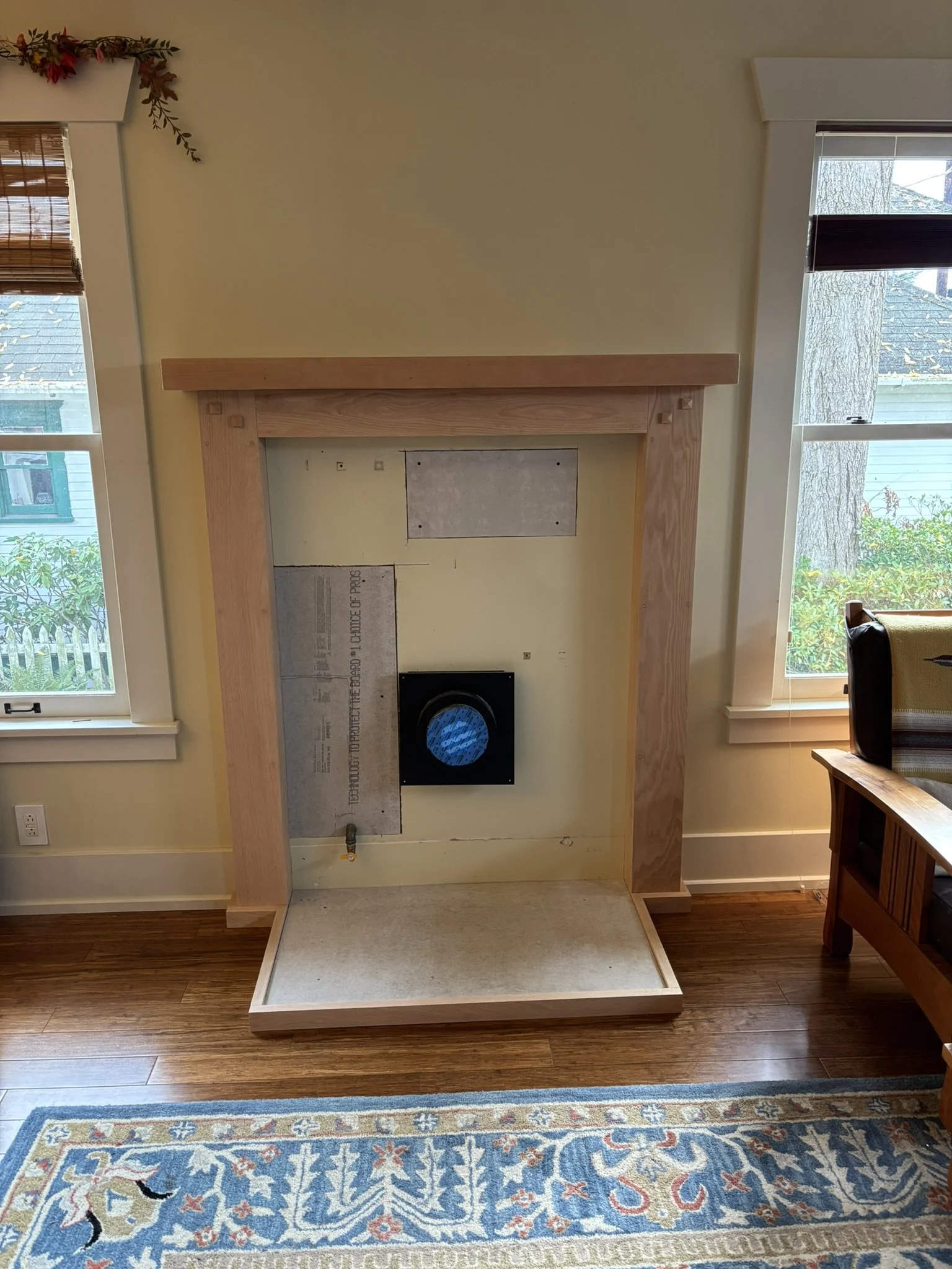 Empty fireplace with a wooden mantel, surrounded by a incomplete yellow wall with insulation panels visible, in a room with hardwood floors and windows on either side.