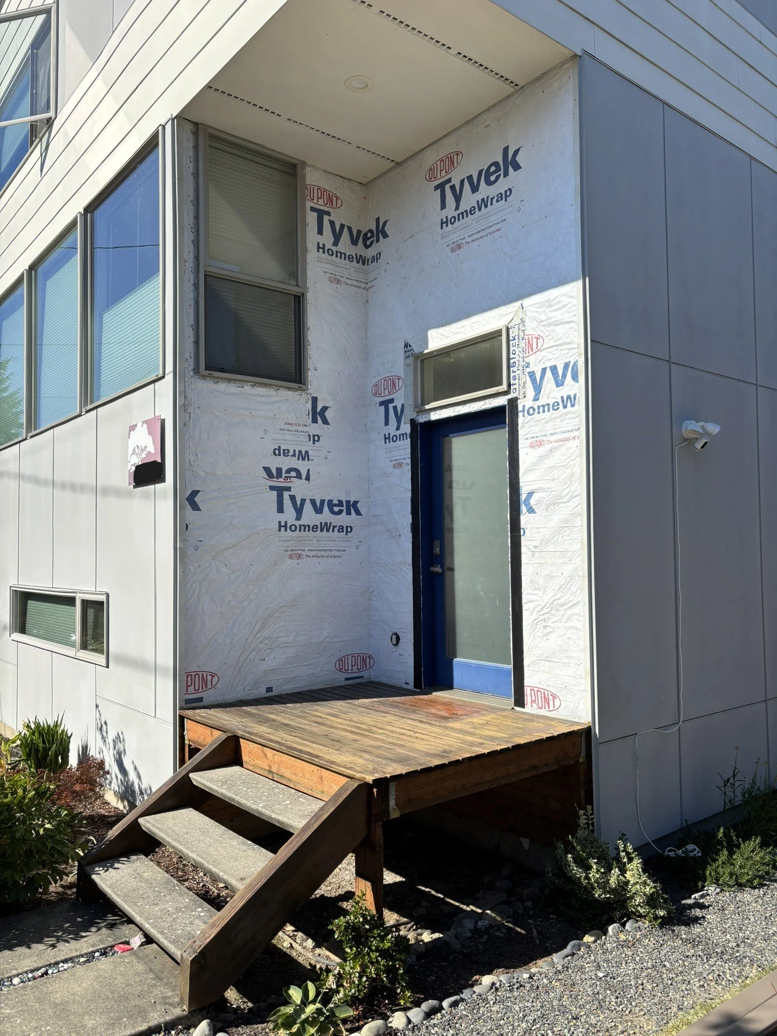Front entrance of a building under construction with a small wooden staircase leading to a glass door, partially covered by Tyvek HomeWrap.